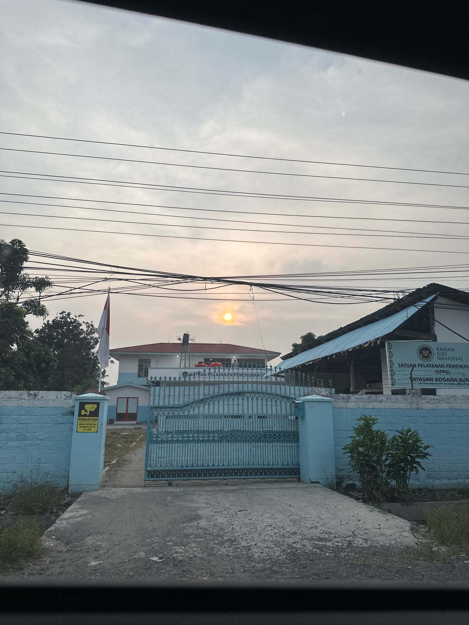 The image shows the exterior of a building with a blue fence and gate. A red and white Indonesian flag is flying on a flagpole. There is a sign on the wall that reads "PERHATIAN AREA DIPANTAU KAMERA CCTV 24 JAM" which translates to "ATTENTION AREA MONITORED BY CCTV 24 HOURS". Another sign on the right side of the image reads "BADAN GIZI NASIONAL SATUAN PELAYANAN PEMENUHAN (SPPG) YAYASAN KOGANA SUMATERA". The sun is setting in the background, casting a warm glow over the scene. The building appears to be a government or official institution. The location is specified as the City of Binjai, Indonesia.