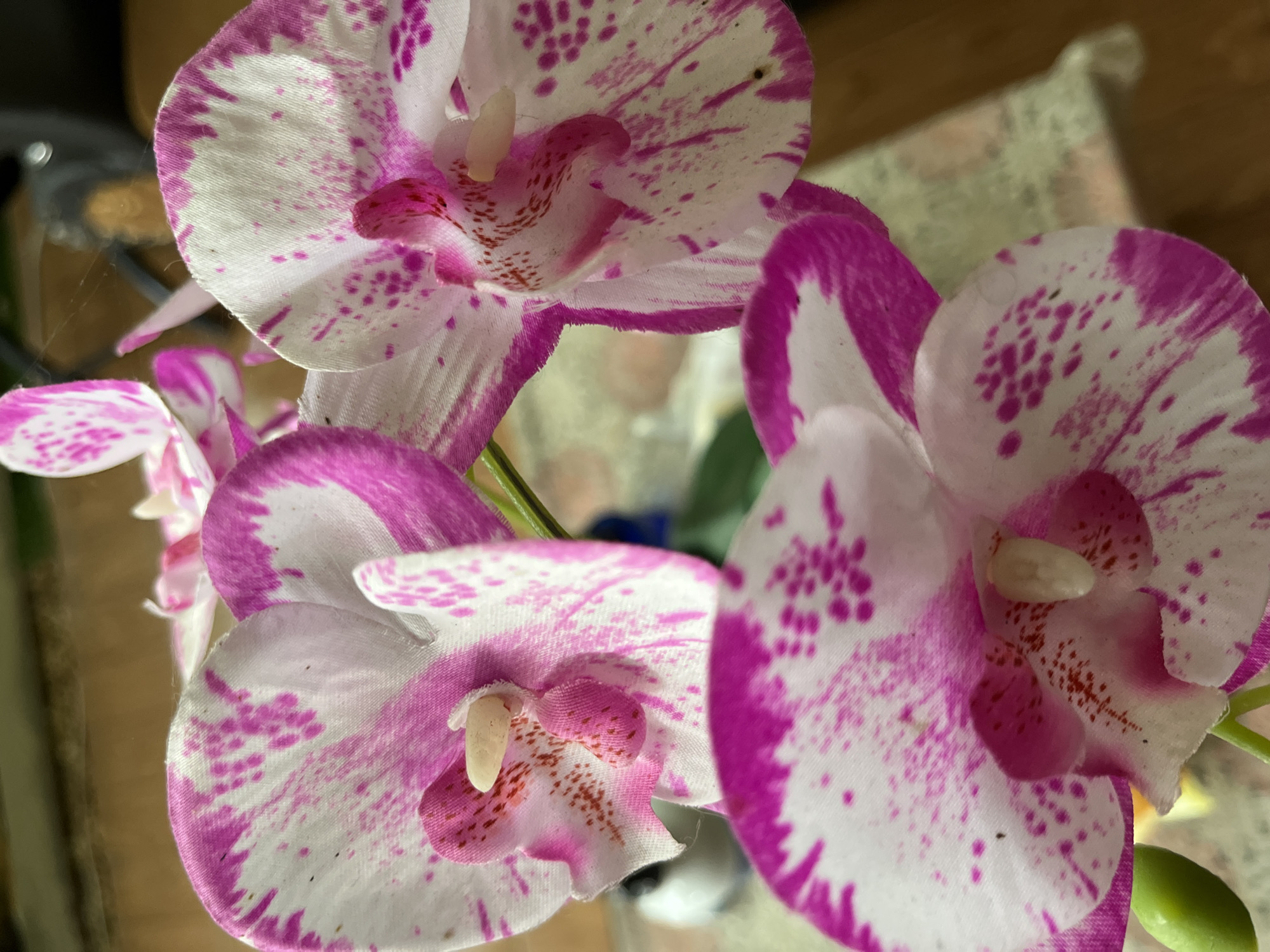 Three artificial orchids with white petals and pink speckles and edges are arranged in a close-up shot. The orchids are in focus, with a blurred background showing a patterned surface and wooden flooring. The scene appears to be indoors. There is no discernible connection to the city of Tasikmalaya, Indonesia.
