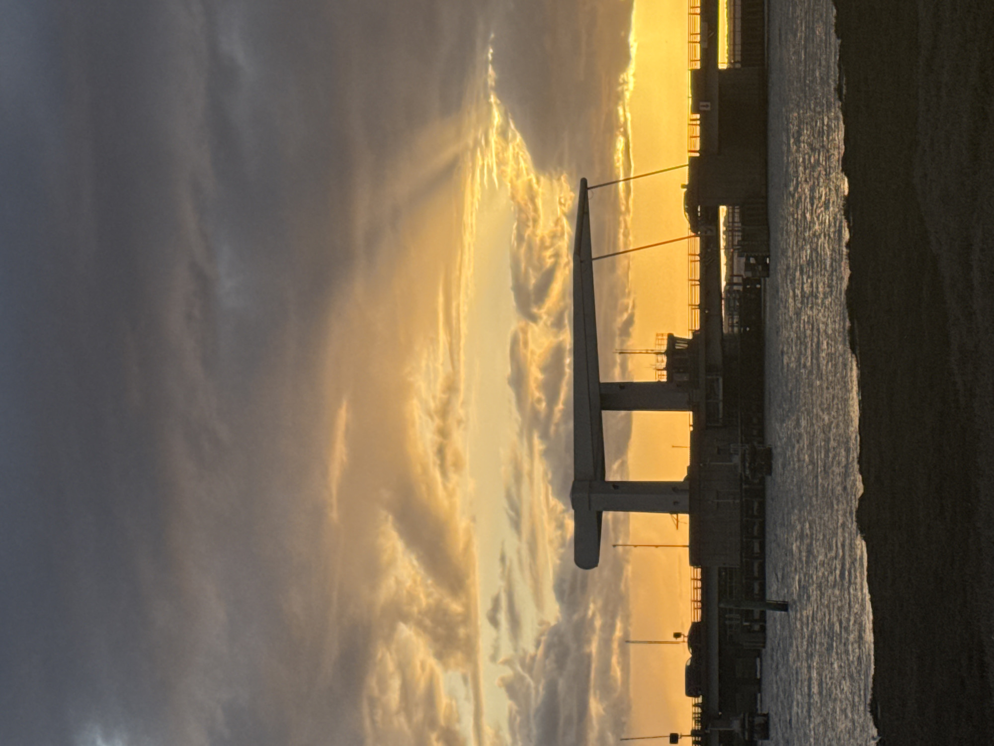 A long, angled shot captures the pier in Great Yarmouth, United Kingdom, set against a vibrant sky at sunset. The pier’s steel structure is prominent in the frame, with the sea visible along the edge, blending into a dark strip. Silhouetted buildings and smaller structures line the pier. The sky displays a dramatic sunset, with golden light piercing through the clouds.