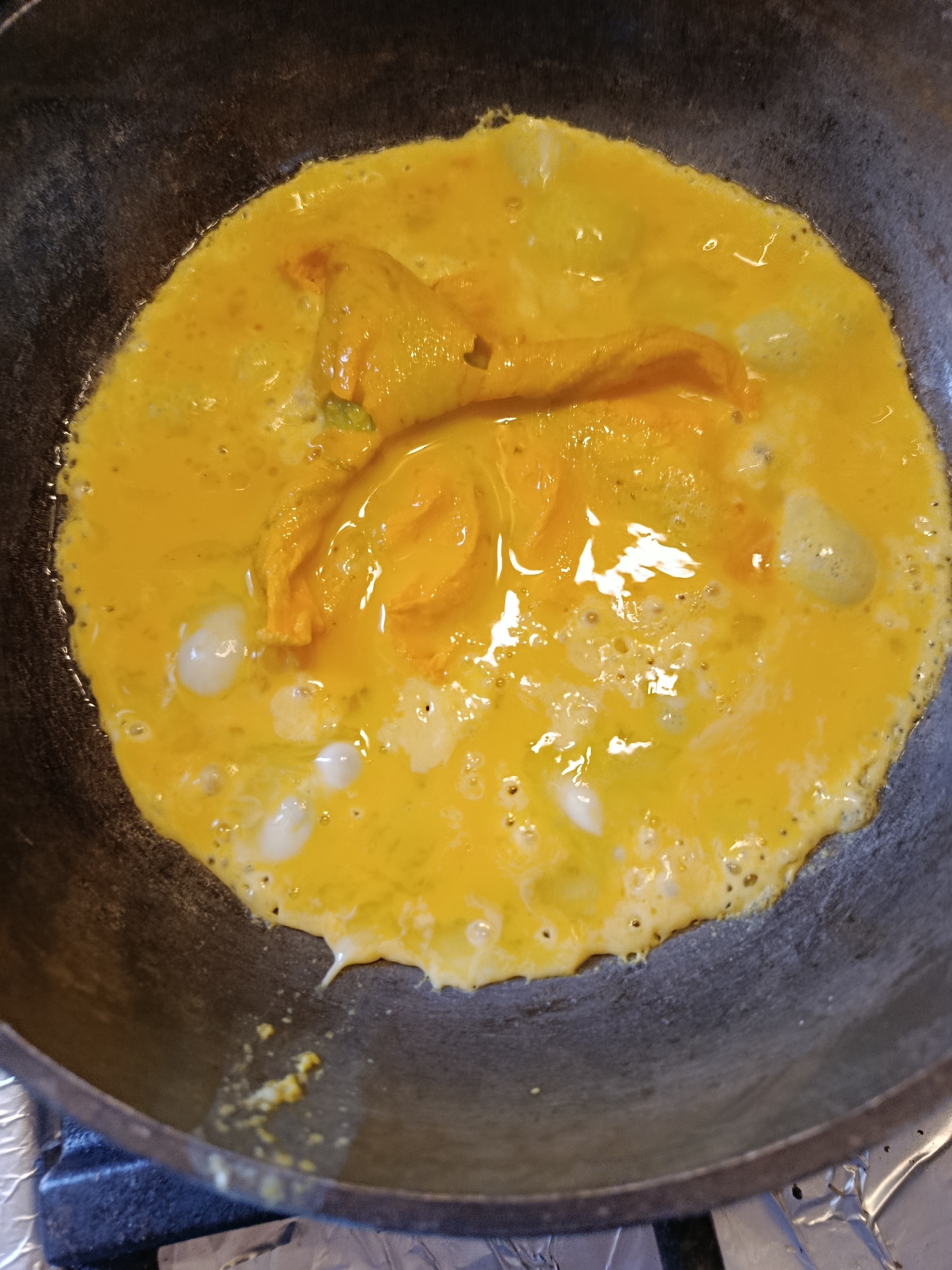 An overhead shot shows a close-up of a dark, cast-iron pan where scrambled eggs are being cooked. The eggs are a bright yellow and have some lighter-colored, opaque sections, suggesting they are partially cooked. There are small bubbles and some areas of liquid on the surface of the eggs. The pan itself has a matte, dark gray finish. The scene appears to be in a kitchen, and the pan is placed on a stovetop which is partially covered with aluminum foil in the lower foreground. The image does not provide any specific details about the location in the city of Верея, Russia.