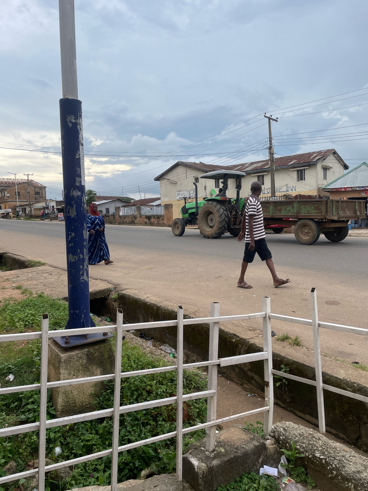 The image depicts a street scene in Jos, Nigeria, under an overcast sky. In the foreground, a blue and white pole stands prominently on the left. A woman in a blue and white striped robe walks on the sidewalk to the left of the pole. Further down the street, a man wearing a black and white striped t-shirt and black shorts walks across the road. Behind him, a green tractor with a trailer is moving along the road. Several buildings with corrugated iron roofs line the street. A white metal fence runs along the bottom of the frame, separating the sidewalk from a drainage ditch filled with greenery and debris.