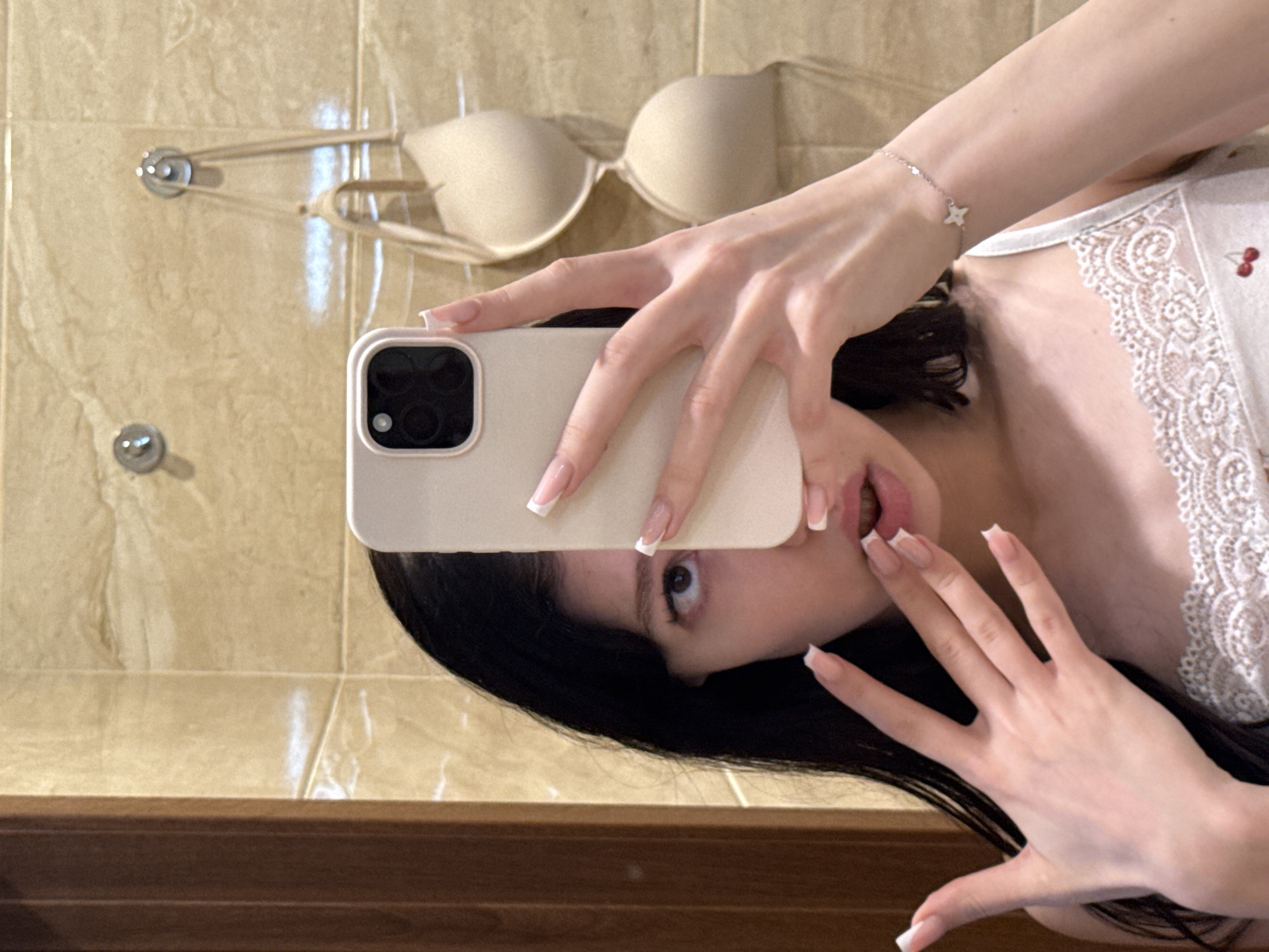 A young woman with long, dark hair is taking a mirror selfie in what appears to be a bathroom. She is holding a light-colored smartphone in her right hand, partially obscuring her face. Her left hand is raised near her face, with her fingers spread. She is wearing a white camisole with lace trim and a small cherry pattern. In the background, beige tiled walls are visible, with a bra hanging from a hook. To the left of the frame, a portion of a wooden door or cabinet is visible. The lighting suggests an indoor setting, likely a domestic environment. No specific landmarks or identifiable features place the location in Khimki, Russia.