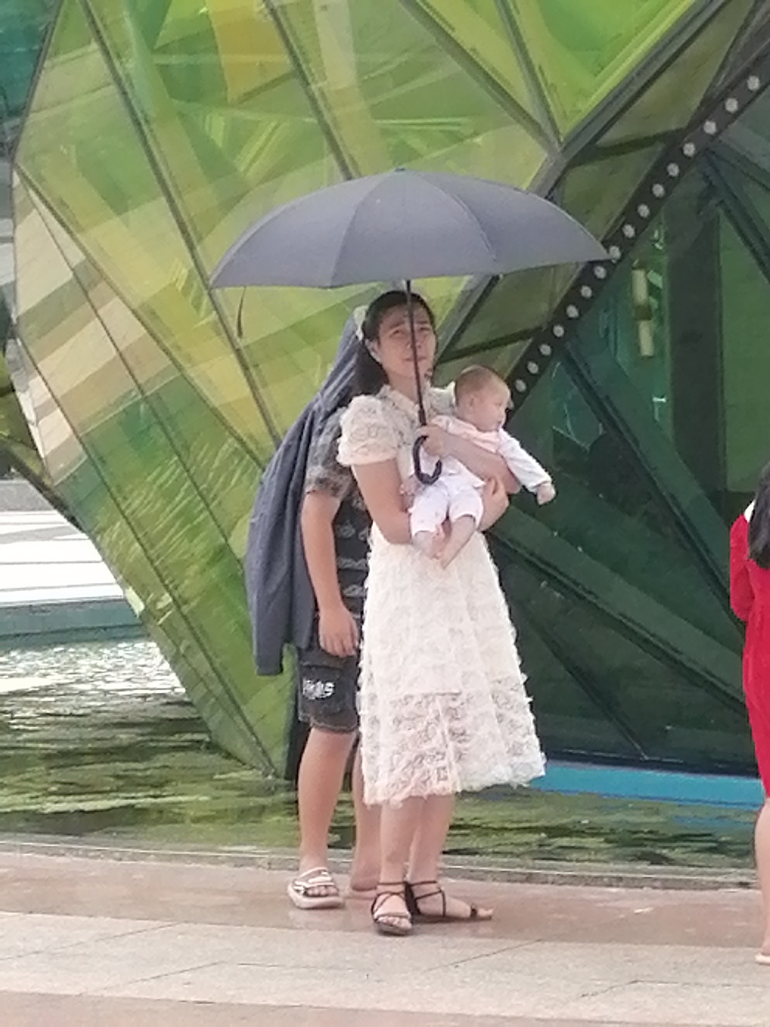 The image depicts a woman holding a baby under a black umbrella. The woman is wearing a white lace dress and the baby is dressed in white. Behind the woman, a person in dark clothing, possibly a jacket draped over their shoulder, is partially visible. They are wearing dark shorts with a white design. The scene appears to be outdoors, with a large, abstract structure with green, translucent panels in the background. There is a body of water in the lower left portion of the image. A person in a red garment is visible on the far right. The ground is paved with light-colored tiles. The overall impression is a candid moment captured in a public space. It is not possible to determine the specific location in Da Lat, Vietnam, from the provided image.