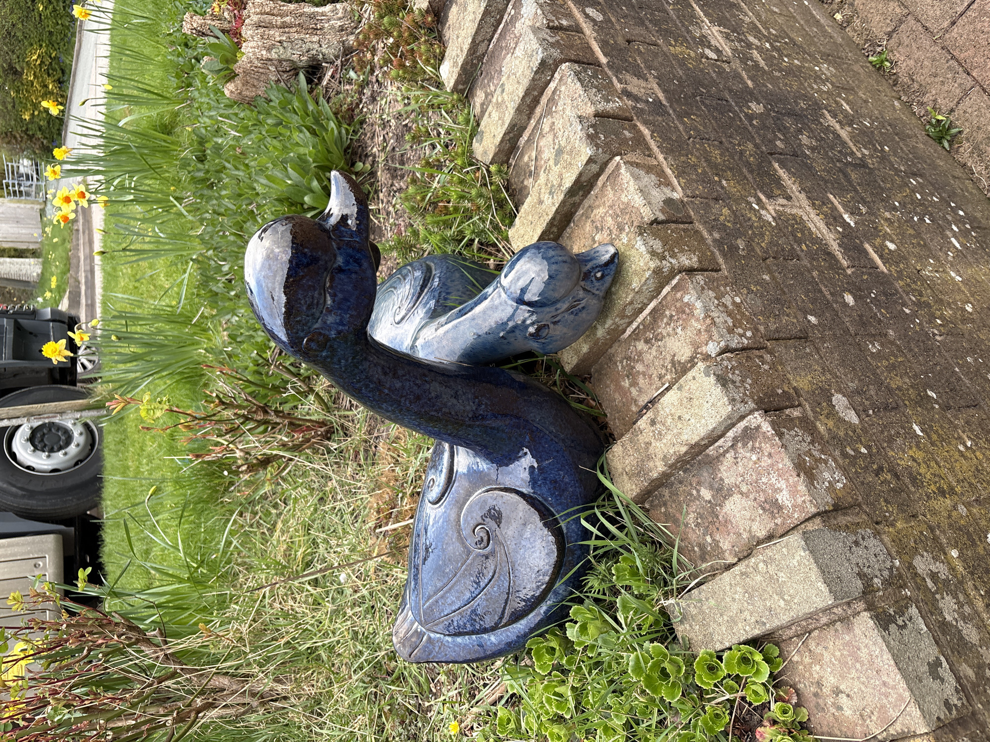A ceramic blue heron sculpture stands in a garden bed next to a brick retaining wall. The sculpture is shiny and detailed, with a long neck and head raised upwards, and a shell-like base adorned with a spiral design. The garden bed contains grass and other greenery. Yellow daffodils are in the background. The location is Shrewsbury, United Kingdom. A portion of a white vehicle is visible to the left.