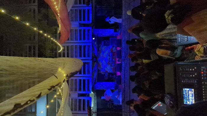An outdoor evening event is depicted in Dhaka, Bangladesh, likely at an open-air venue or rooftop. The scene is illuminated by decorative string lights and stage lighting, casting a predominant blue hue on the stage area.
**Environment:** Overhead, multiple large, off-white fabric drapes, some featuring dark patterns, are suspended, crisscrossed by strands of warm-toned fairy lights. A red fabric drape is visible on the right. In the background, behind the stage, a building facade with several horizontal, slatted rectangular openings is present. The sky is dark, indicating night.
**Stage:** A raised stage occupies the mid-ground, featuring musical equipment including drums, keyboards, and multiple floor monitors or speakers arranged along the front edge. The stage backdrop is decorated with abstract, patterned art. Two stylized, flat cut-out figures stand on the stage: one on the left, colored red and blue, depicts a person playing a percussion instrument; another on the right, white, depicts a person holding a microphone.
**Audience:** A dense crowd of people occupies the lower portion of the image, seated or standing on the ground directly in front of the stage. The individuals primarily have dark hair, and their attire varies, including light-colored and dark garments. They are oriented towards the stage, observing the performance.
**Foreground:** In the immediate foreground, two professional audio mixing consoles are visible. The console on the left features an illuminated digital screen displaying graphical information, along with multiple faders and controls. A laptop computer is positioned near this console. The presence of these consoles suggests a technical control area for the event.