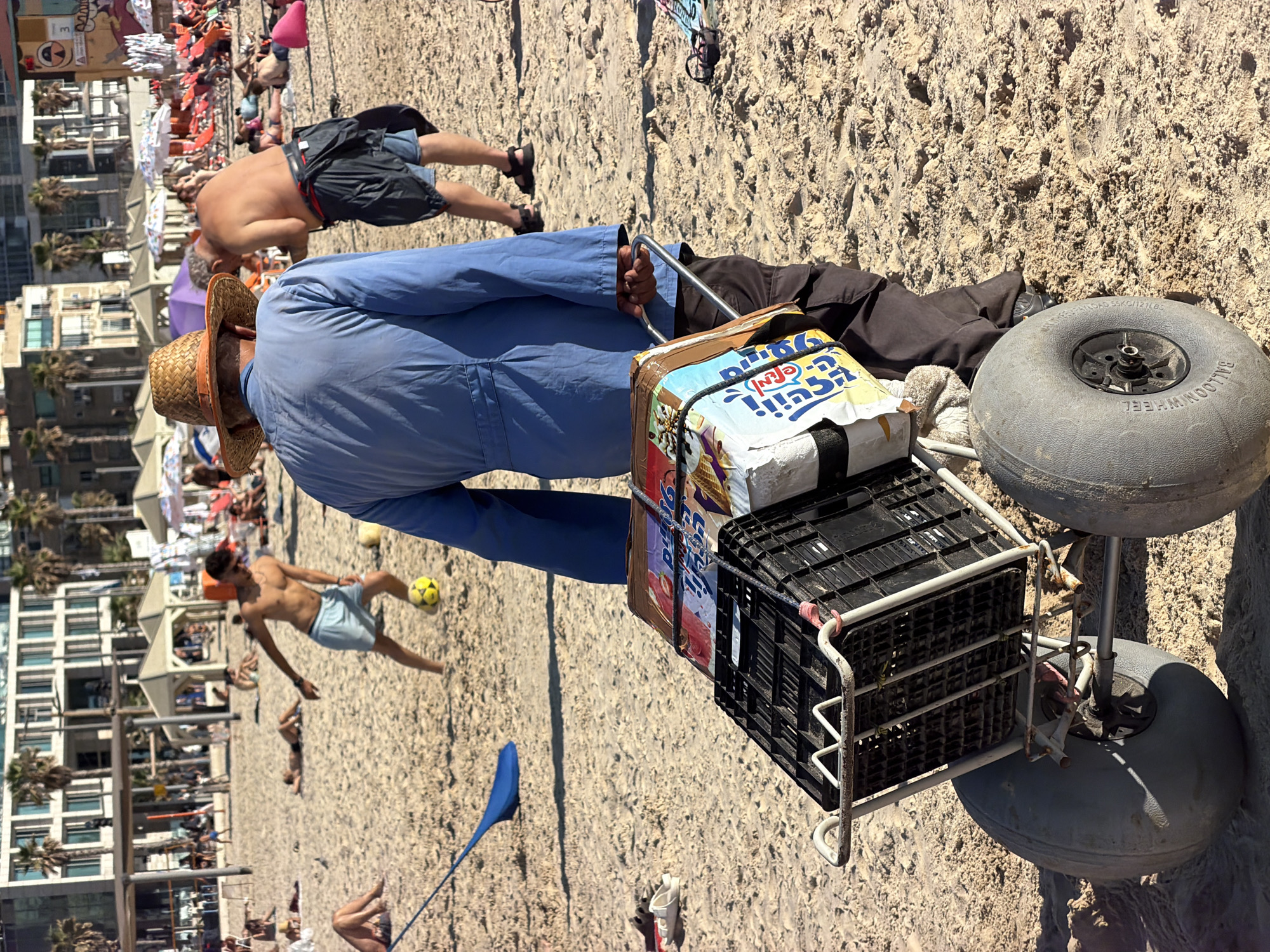 The image depicts a busy beach scene in Tel Aviv, Israel, on a sunny day. The primary subjects are a person in a blue jumpsuit and straw hat pushing a cart filled with items, and several other beachgoers scattered across the sand. The cart has a large, grey balloon wheel, designed for sand, and is loaded with a black crate containing what appears to be packaged food, possibly ice cream, given the visible text and imagery on the packaging.
In the background, numerous people are seen sunbathing, walking, and relaxing. Some are under umbrellas, while others are directly exposed to the sun. Buildings, likely hotels or apartments, are visible behind the beach, indicating an urban coastal setting. Palm trees are also scattered throughout the scene. A young man is playing soccer with a yellow ball in the mid-ground, adding to the lively atmosphere of the beach.
The time of day appears to be midday, suggested by the bright sunlight and the shadows cast by the people and objects. The weather is clearly warm and sunny, typical for a beach day in Tel Aviv. The overall mood is one of leisure and recreation.
Visible text on the packaging includes Hebrew characters, with "טעם" (taste) and "שוקו" (chocolate) being partially legible. The balloon wheel has the text "BALLOON WHEEL" printed on it. The image captures a typical summer day at a popular Israeli beach.