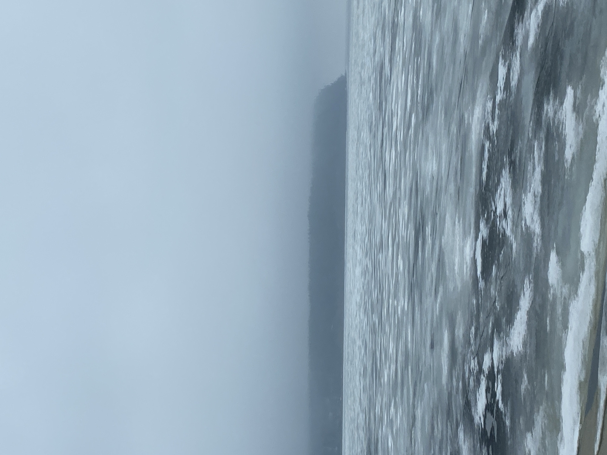 The image depicts a landscape in Chaikovsky, Russia, characterized by a body of water partially covered with ice. The right side of the image features the icy surface, showing patterns of ice and some snow. A hazy or foggy atmosphere is present, obscuring the horizon and diminishing visibility. On the left, a muted sky or fog dominates, creating a monochromatic palette. A vague silhouette of a shoreline or landmass is seen in the middle ground.