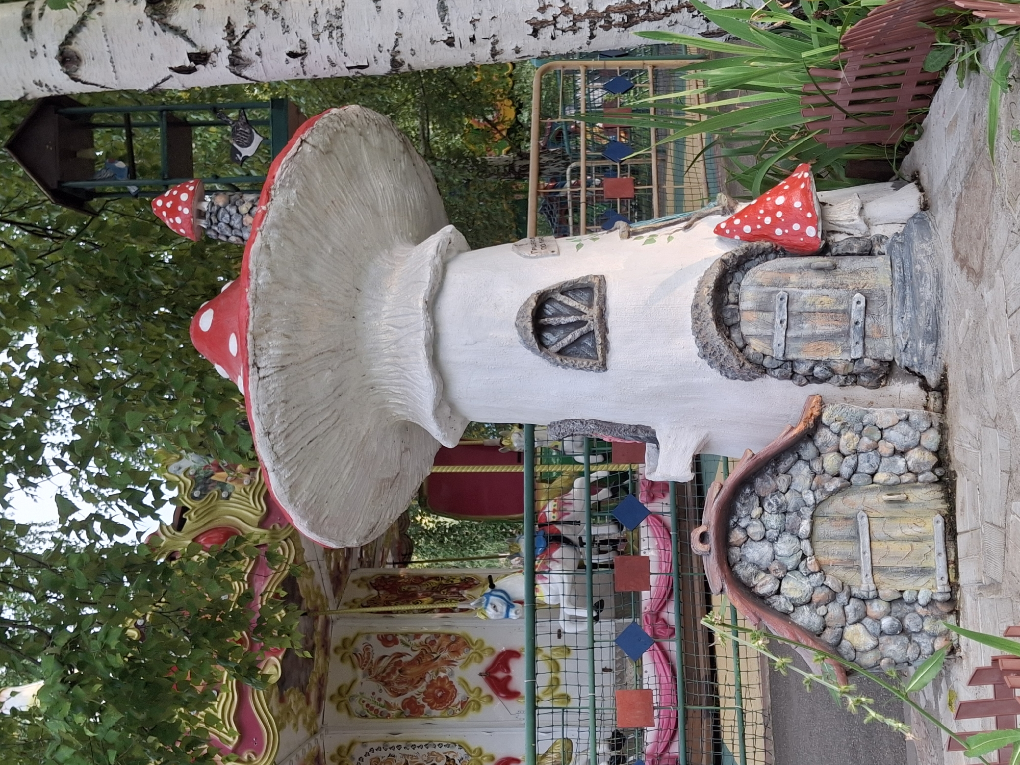 The image depicts a whimsical scene in the city of Chaikovsky, Russia, featuring a carousel and a fairy-tale mushroom house. In the background, a portion of a brightly colored carousel with ornate decorations and painted horses is visible. In the foreground, a large, white mushroom-shaped house with a red, spotted cap serves as the central subject. Attached to the mushroom house is a smaller, stone-clad structure with a wooden door, designed to resemble a miniature hobbit-hole. Another small, red-capped mushroom adorns the side of the main mushroom house. A bird feeder made of dark wood is mounted on a birch tree to the right of the mushroom house. The ground is paved with stone. Lush green foliage and a wooden fence complete the enchanted setting.