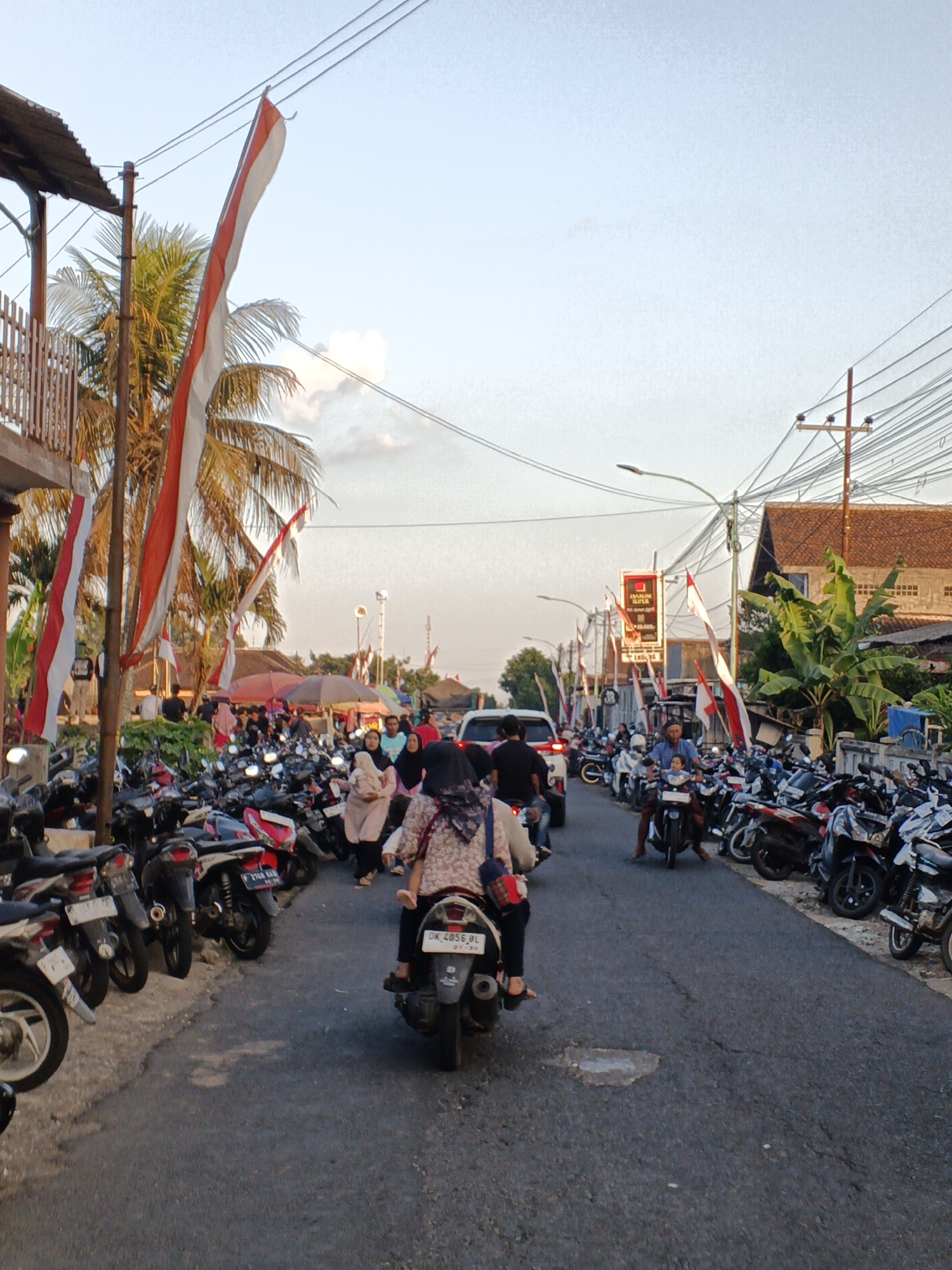 The image depicts a street scene in Balak, Indonesia, likely during daytime with a clear sky and warm lighting. The street is lined with numerous parked motorcycles on both sides, indicating a busy area. Indonesian flags, red and white, are prominently displayed, hung vertically on poles and wires, suggesting a national celebration or event. A few people are seen riding motorcycles, with one in the foreground carrying a passenger. Others are walking or standing near the parked vehicles. Buildings with traditional tiled roofs are visible on the right side of the street, along with some trees, including palm trees. Electrical wires and streetlights are also part of the urban environment. The road surface appears to be asphalt with some wear and tear. The overall atmosphere suggests a lively community gathering or marketplace.