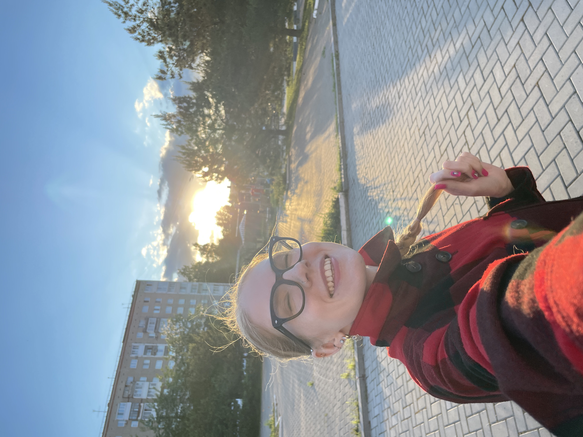This image is a cheerful outdoor selfie of a young woman, taken in Izhevsk, Russia, likely during late afternoon or early evening. The photograph is currently rotated 90 degrees counter-clockwise.
The main subject is a blonde woman with a wide, happy smile, showing her teeth. She wears black-rimmed glasses and a red and black plaid shirt. Her hair appears windswept, and she is playfully holding a section of her braided hair with her right hand. Her fingernails are painted pink with small star embellishments. Several piercings are visible on her left ear and nose, with a distinct earring dangling from her left ear.
The background reveals an urban or suburban environment. A multi-story, light-colored residential apartment building, consistent with Soviet-era architecture, is visible on the left side of the frame, partially framed by trees. A paved path or sidewalk, made of light-colored interlocking pavers, extends into the distance on the right. The sun is low in the sky, positioned behind the subject, casting a warm, golden glow and creating sun rays and lens flares. The sky is mostly clear with some scattered clouds. The overall mood is bright and joyful. No visible text is present in the image.