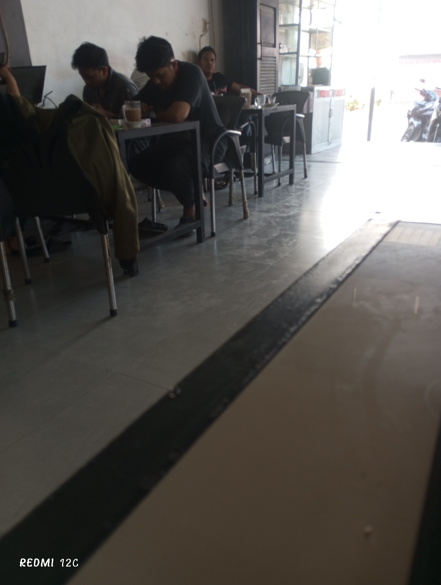 The image depicts the interior of a cafe or coffee shop in Lhokseumawe, Indonesia. Three men are seated at tables, appearing to be engaged with their mobile devices. A cup of coffee is visible on the table. The floor has a dark, contrasting stripe running along its surface. Natural light streams in from an open area, where parked motorcycles are visible.