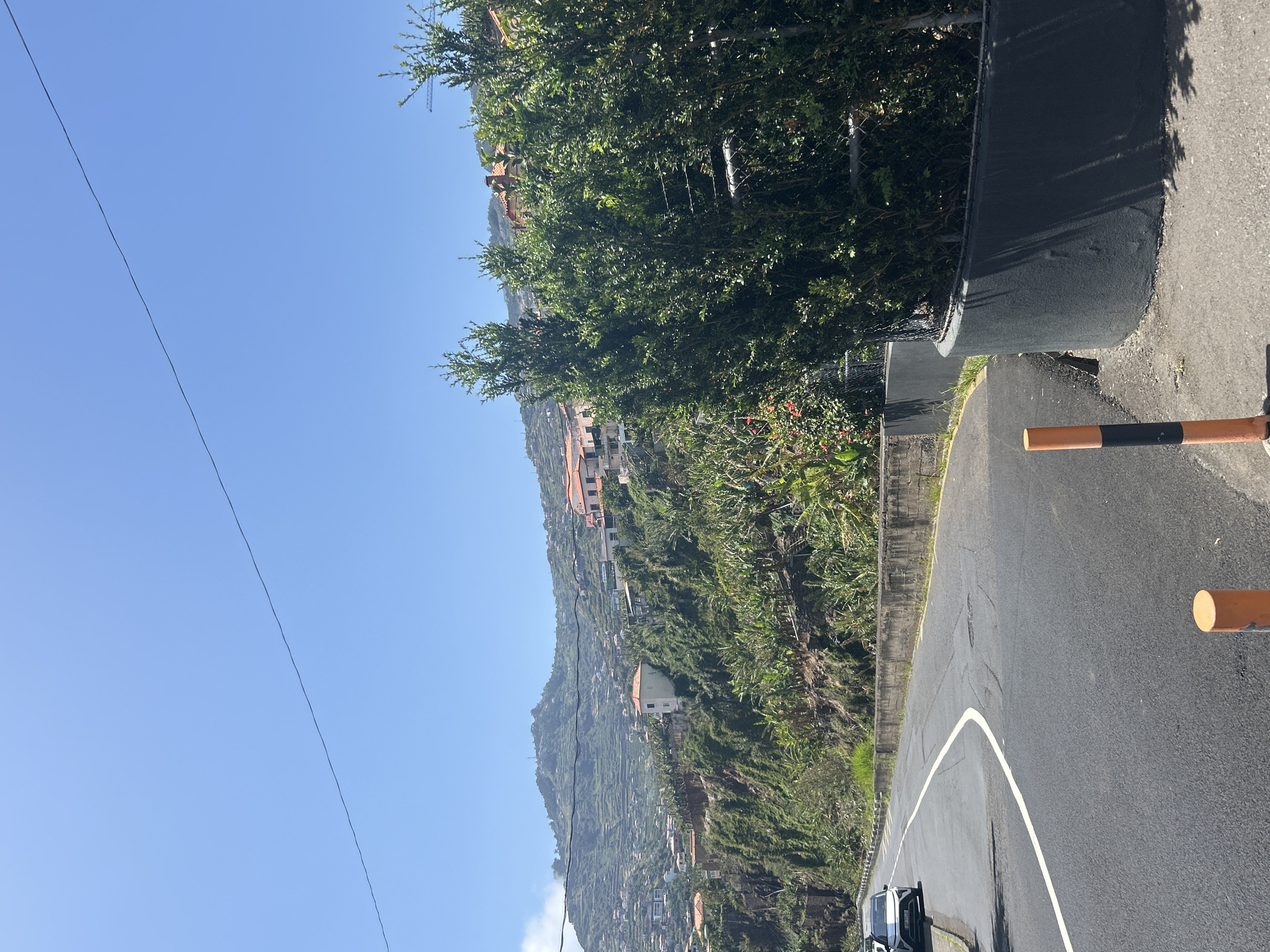 The image depicts a street scene in Câmara de Lobos, Portugal. A paved road with a white lane marking curves through a landscape of terraced hillsides covered in lush vegetation. Several buildings, including houses with red tile roofs, are visible on the hillside. The sky is clear and blue. A portion of a barrier painted with orange and black stripes is seen on the right side of the road. A power line is visible in the upper left part of the image.