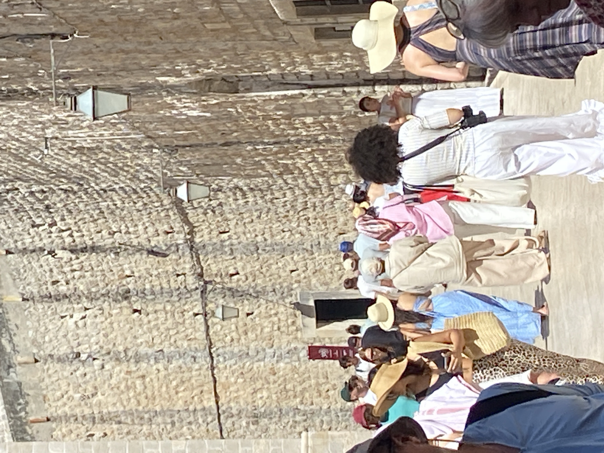 In Dubrovnik, Croatia, on a bright sunny day, a group of tourists walks along a narrow cobblestone street lined with ancient stone walls. The walls, constructed from rough-hewn, light-colored stones, show signs of age and weathering, with visible mortar in between. Several decorative, old-fashioned street lamps are mounted on brackets attached to the wall at intervals, casting shadows on the textured surface.
The street is populated by a diverse group of people, predominantly dressed in casual, summery attire. Many wear hats, including wide-brimmed straw hats and baseball caps, for sun protection. Some individuals are carrying bags, such as straw totes and backpacks, suggesting they are exploring the city. The people are walking in the same direction, appearing to be part of a tour group or casually strolling together. Clothing styles range from light-colored linen outfits to patterned skirts and dresses. One person in the foreground, viewed from the back, has dark curly hair and is wearing a white pinstripe top and loose white pants, with a camera bag slung over their shoulder. Another woman, also viewed from behind, wears a wide-brimmed straw hat and a patterned dress.
In the middle ground, a doorway is visible, recessed into the stone wall, with a dark door and a small red sign hanging to its left. The overall atmosphere is one of a popular tourist destination, with people enjoying the historical ambiance and architecture. The sunlight creates strong contrasts between illuminated areas and shadows, highlighting the rugged texture of the stone walls.