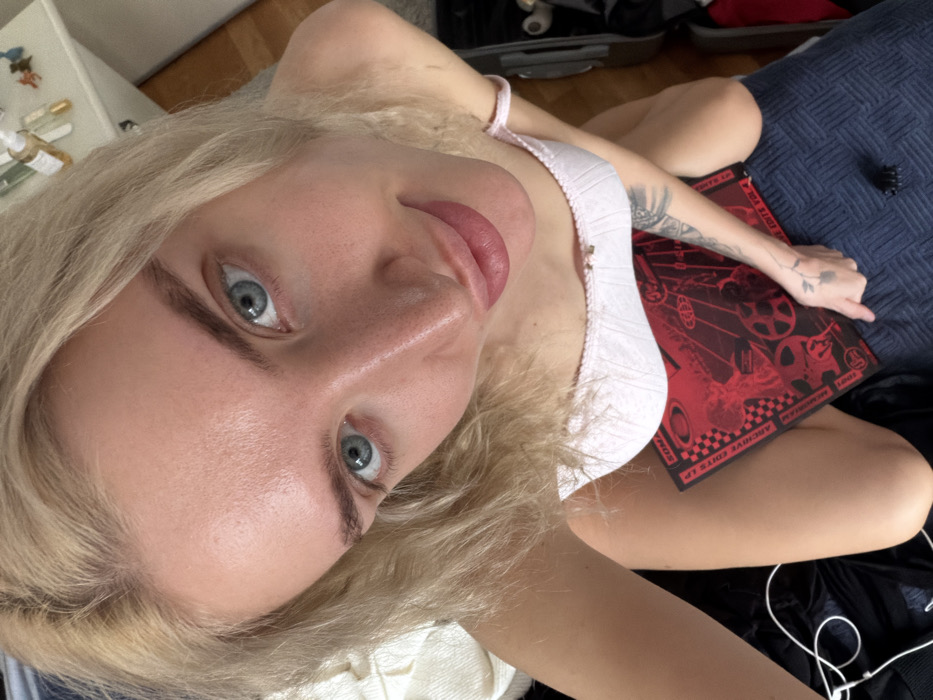 A female individual with blonde hair, blue eyes, and fair skin is depicted in a high-angle indoor photograph taken in Budva, Montenegro. She is looking directly at the camera with a slight smile. The subject is wearing a light-colored bralette or camisole top. A tattoo featuring a floral motif is visible on her left forearm. A red album cover or book with black graphic designs and text, including "SOMA" and "ARCHIVE EDITS VOL. 4," rests on her lap. Her right hand is positioned on the dark blue patterned fabric, possibly bedding or upholstery, near the red object. A black hair claw clip is present on the blue fabric to her right. In the background, a white bedside table with several small bottles is visible on the right side, and a white power cord is visible on a dark surface to the left.