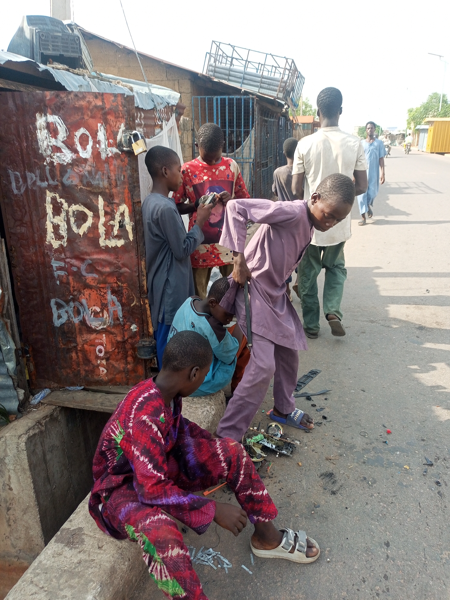 A daytime outdoor shot depicts several individuals in what appears to be an urban setting in an unidentified city. At the forefront, a child wearing a patterned red and purple outfit is seated on a cement structure. To the child's left, there is a rusted corrugated metal wall with "BOLA" and other unreadable text painted in white. Several children are grouped near this wall, with one wearing a red patterned shirt holding an electronic device, and another in a blue shirt seated on the ground. A boy in a purple outfit bends over, seeming to interact with broken electronic components scattered on the asphalt street. In the background, two men are walking on the street away from the camera. Various buildings are visible in the background, including structures with corrugated metal roofs. The environment suggests a lower-income area.