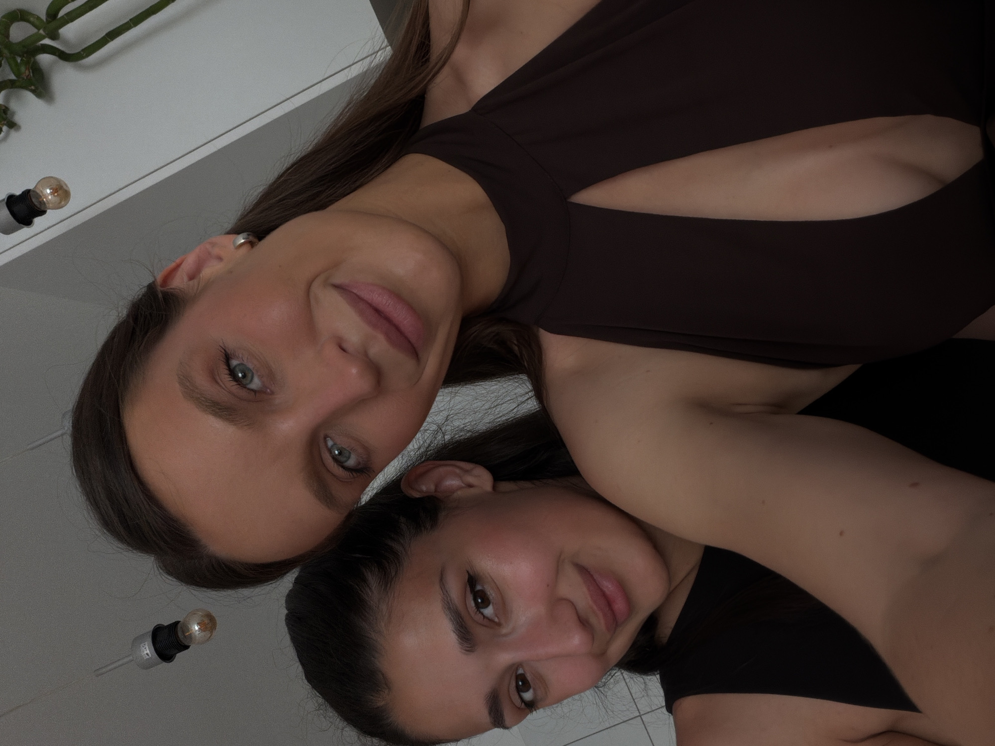 A close-up, selfie-style indoor portrait features two young adult females positioned closely together in a residence located in Budva, Montenegro. The woman on the right, the primary subject, has long, dark brown hair and light blue-green eyes. She wears a dark brown, halter-neck top with a keyhole cutout centered on the chest, revealing cleavage. Her expression is a gentle, closed-mouth smile, and a small, silver-toned hoop earring is visible on her left ear. Her left arm and shoulder are also visible.
Partially obscured behind and to the left of the primary subject is another woman, with dark hair and dark brown eyes, exhibiting a similar gentle smile. She wears a black sleeveless top, and her right arm is visible.
The background shows interior details: a light-colored wall, a single bare light bulb suspended from the ceiling by a grey cord in the upper left, and a multi-stemmed green plant (possibly bamboo) in the upper right. The lighting appears to be soft and warm, characteristic of indoor ambient light.
