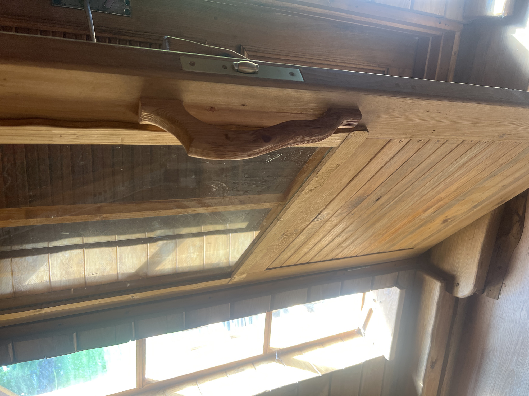 The image shows a rustic wooden door with a large, hand-carved wooden handle. The door is partially open, revealing a glimpse of a bright interior with wooden beams and possibly a window. The door itself is constructed from horizontal wooden planks, with a smaller section of vertical planks above a glass pane. A metal latch mechanism is visible on the door frame. The flooring appears to be light-colored wood. The overall aesthetic suggests a cozy, natural setting, possibly a cabin or a rural dwelling in Daulėnai, Lithuania, given the context. No people are present in the image.