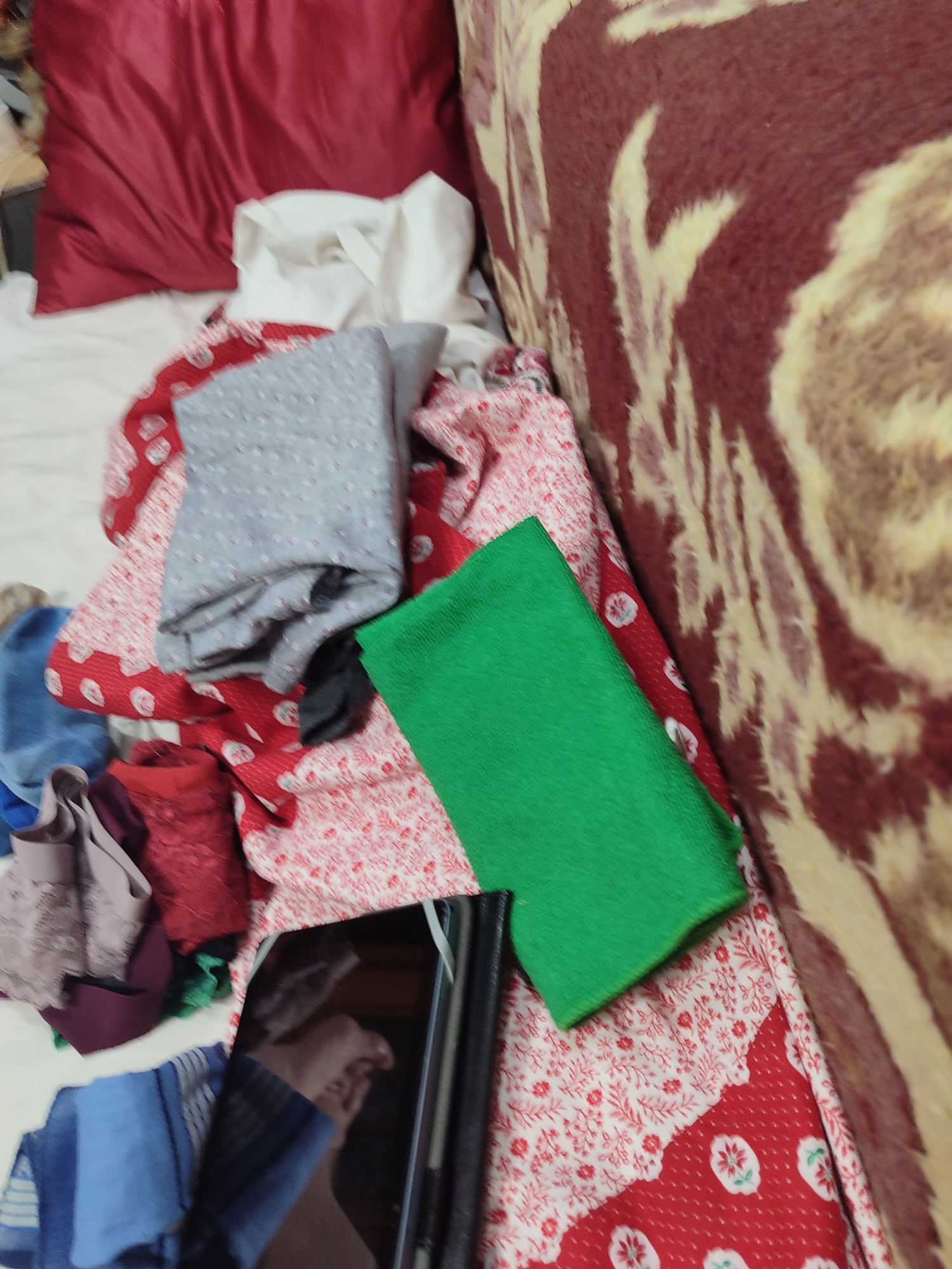 The image shows a pile of fabric scraps on a bed. The fabrics are of various colors and patterns, including red with white floral prints, grey with small white dots, and a solid green fabric. There are also some darker colored fabrics, possibly burgundy and navy blue. A black tablet or phone is lying face down on top of the fabrics. The setting appears to be a bedroom, with a red pillow and a patterned blanket visible in the background. The lighting suggests it could be daytime, but there are no specific cues about the weather or time of day. There is no visible text in the image. The overall impression is one of disarray, possibly indicating a crafting project in progress or materials that have been gathered for use. There are no people directly visible, though a reflection of a person's face and hand can be seen on the screen of the tablet. The context of Mykolaiv, Ukraine, is provided but not visually evident in the image.