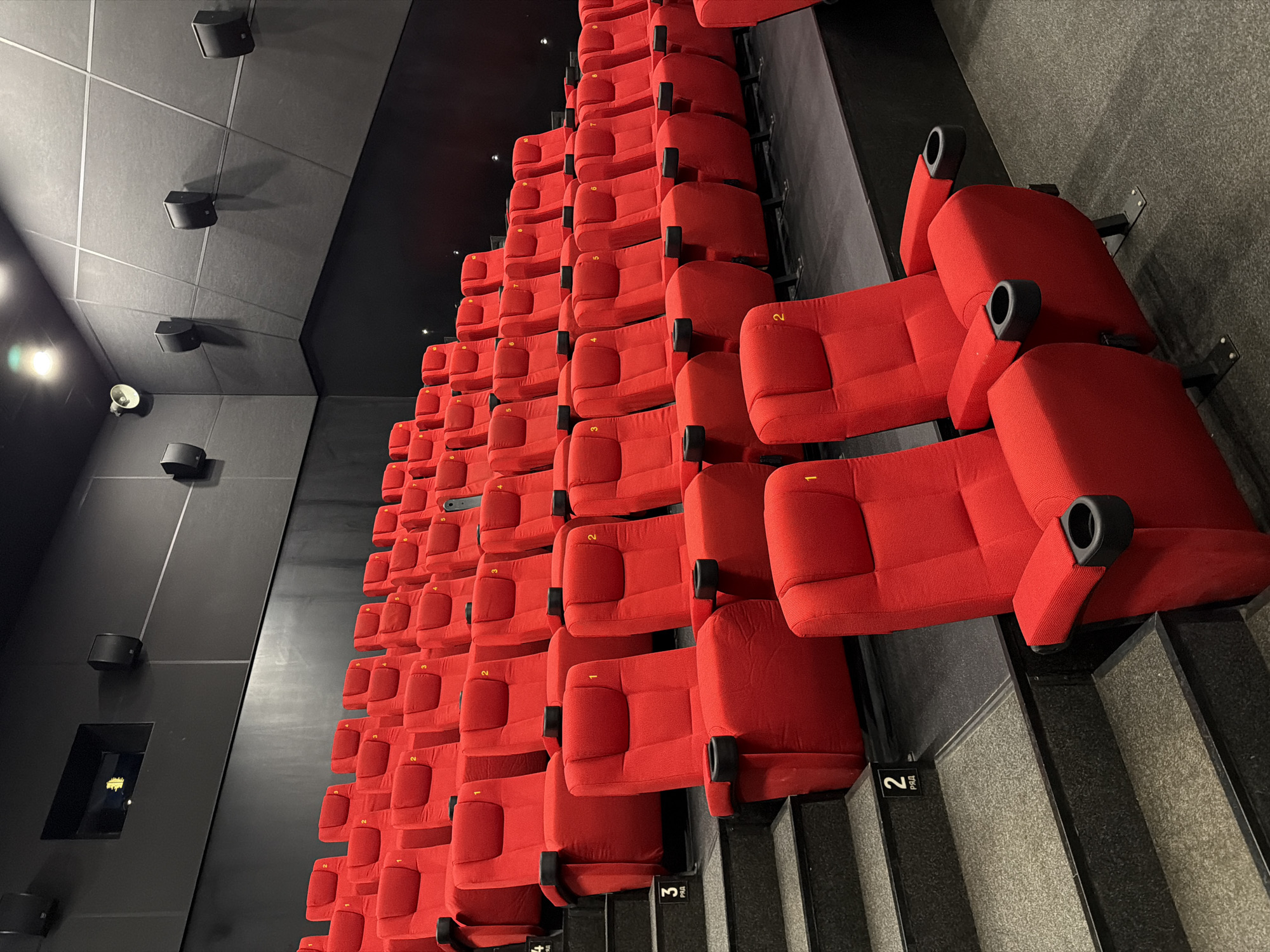 The image shows a section of a movie theater, likely in Unknown, Unknown, as there is no location information provided. The scene is an overhead shot of rows of red seats in a darkened theater. The seats are upholstered, with armrests and cup holders. Each row is slightly elevated compared to the row in front of it. The theater walls and ceiling are dark gray to black with mounted speakers along the walls. Steps lead up to the rows of seating and include row numbers. There are no people visible in the image.