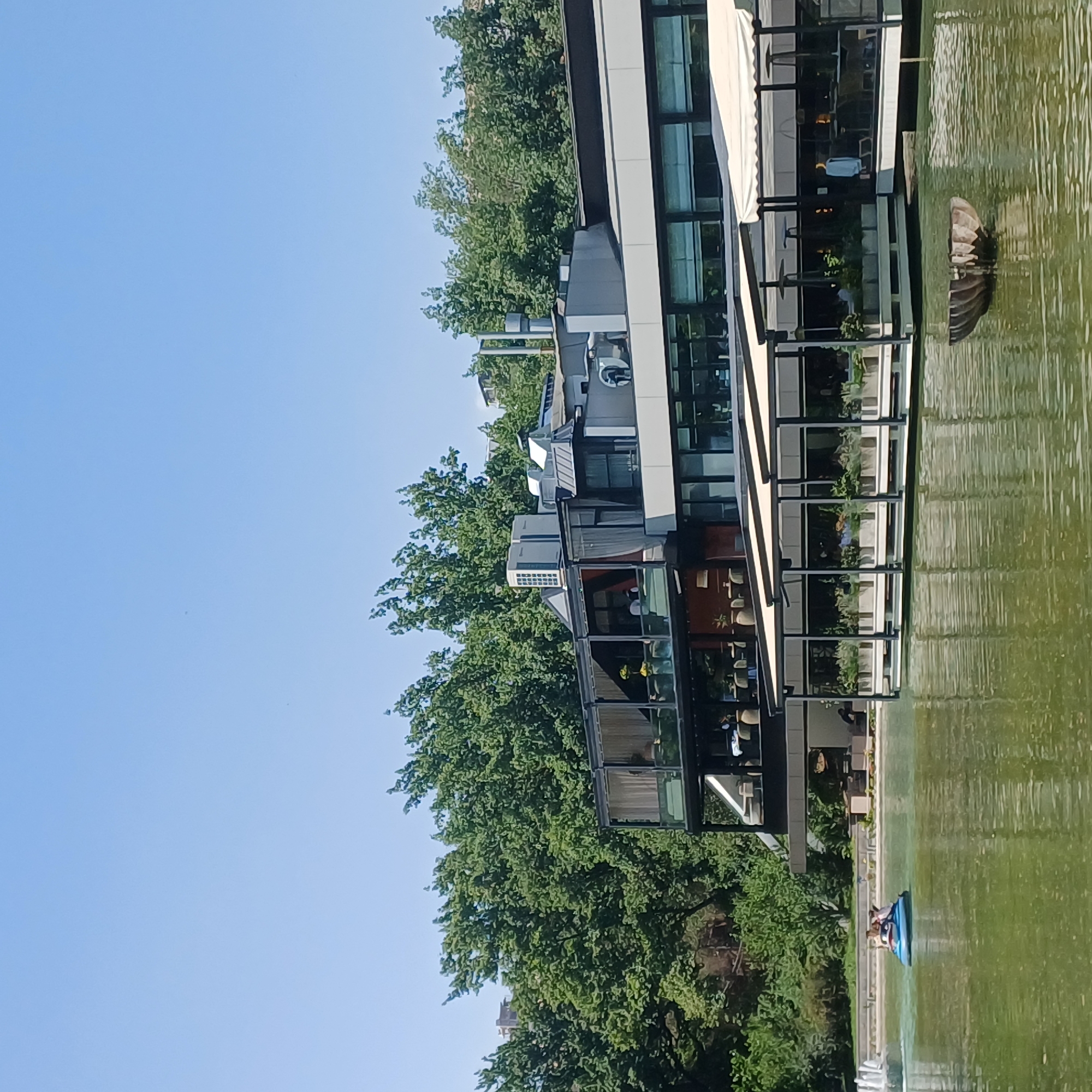 A wide-angle, eye-level shot captures a modern building with multiple levels on the edge of a lake. The building features large glass windows and a dark gray facade, with awnings extending over outdoor seating areas on the lower level. Lush green trees line the background, contrasting with the clear blue sky. In the foreground, the green water of the lake reflects the surrounding environment. A small blue boat with two people is visible in the left portion of the lake, near a fountain. A rustic, bowl-shaped object floats in the water towards the right side of the frame. The scene appears to be a restaurant or leisure area in a park setting.