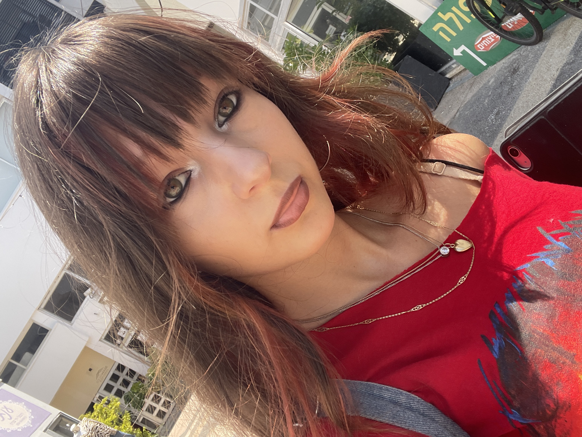 A young woman with brown hair and bangs is taking a selfie outdoors, likely on a street in Raanana, Israel. The background shows a modern building with white and yellow sections, possibly shops, as indicated by a mannequin in a window. A green sign with white text and an arrow points to the right, suggesting directional information or advertisements. The woman is wearing a red top with a graphic design and two gold necklaces, one with a heart-shaped pendant. A denim jacket is visible on her left shoulder. She has heavy eye makeup. A bicycle wheel and part of a bicycle are visible in the lower right corner, near a curb. The lighting suggests it is daytime.