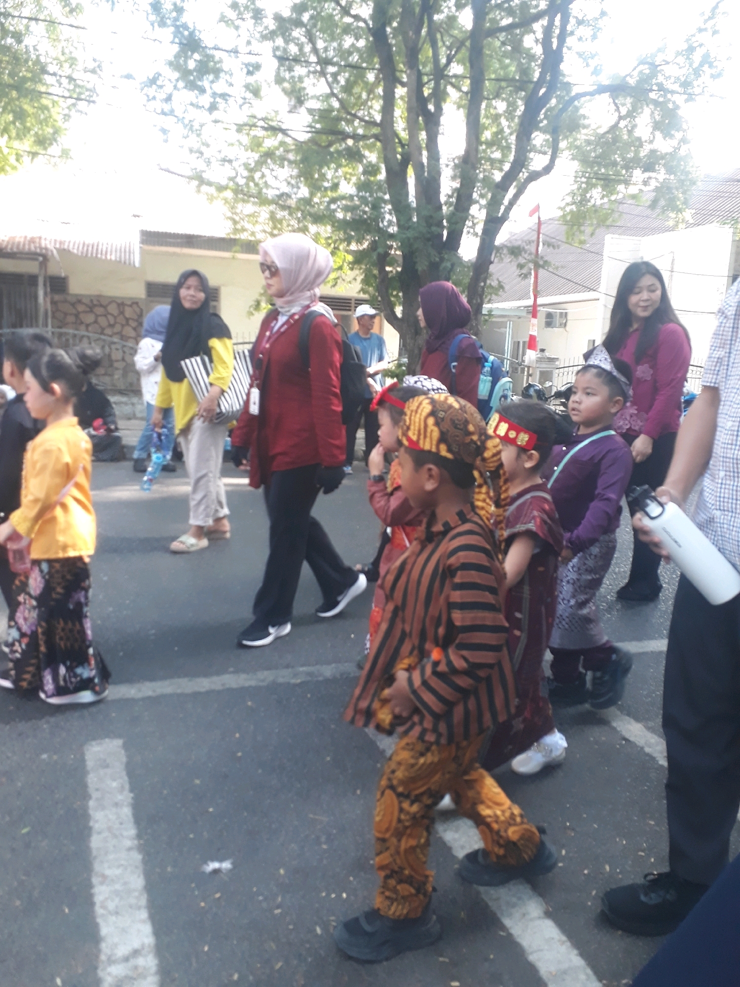 The image shows a group of people, including children and adults, walking on a paved street in what appears to be a procession or parade. The children in the foreground are wearing traditional Indonesian clothing. The street is lined with buildings and trees. One child is wearing a yellow top and a patterned skirt. Another child is wearing a brown and orange striped top and patterned pants. A woman in a red jacket and headscarf is walking behind them, holding a bag. A woman in a purple top is in the background, and another woman in a purple headscarf is walking to her left. The context of Pangkalpinang, Indonesia, suggests this may be a cultural event. The presence of buildings, trees, and a paved street indicates an urban or suburban environment.