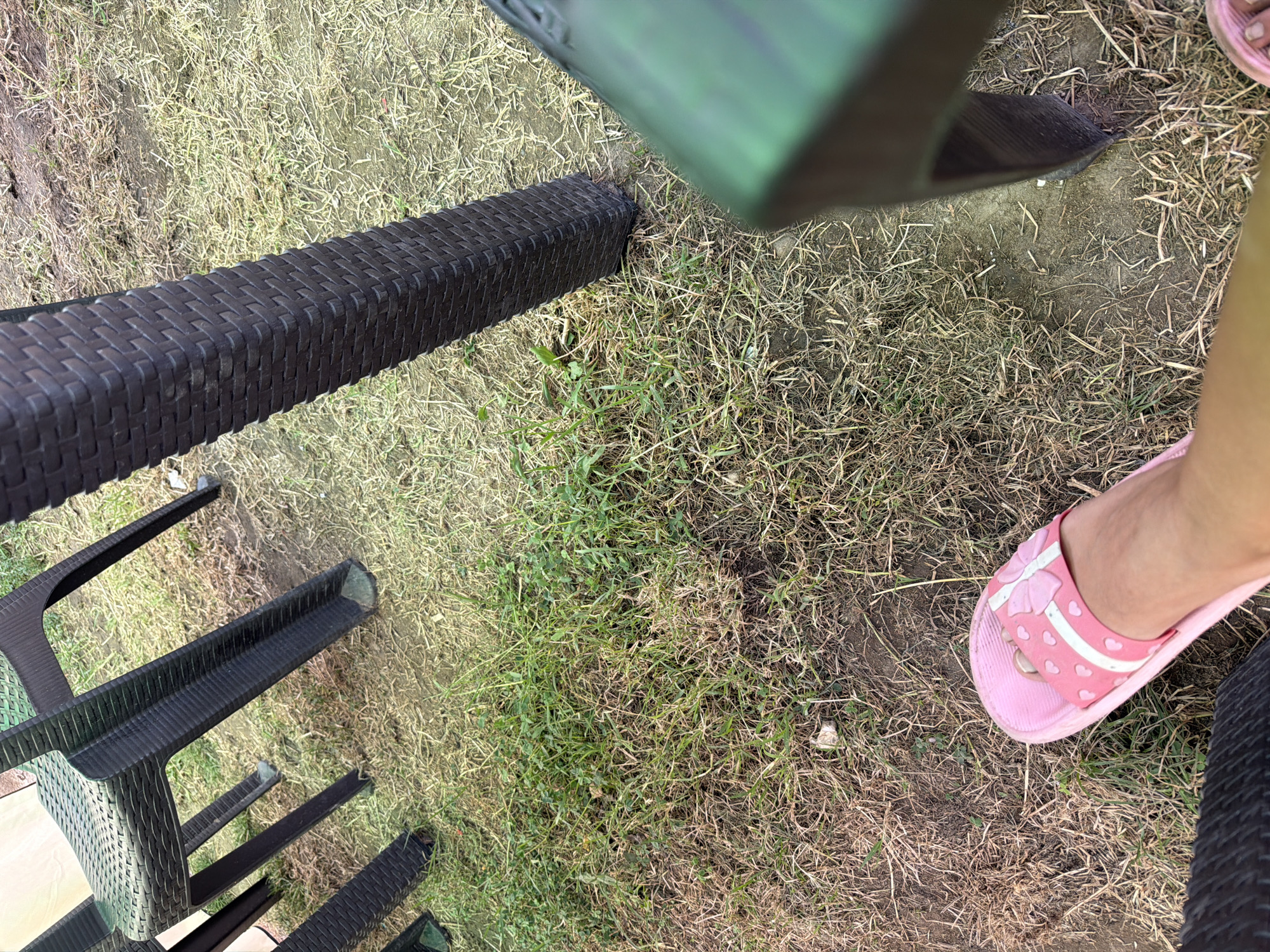 A first-person perspective shot captures a person's foot wearing a pink sandal with a white stripe and a bow, positioned on dry grass and dirt. In the background, a dark brown plastic chair leg is visible, its textured surface resembling woven material. A portion of a green plastic chair is also in the frame, blurred and out of focus. The scene appears to be outdoors, possibly in a park or a casual seating area. The location is Razhevo Konare, Bulgaria.