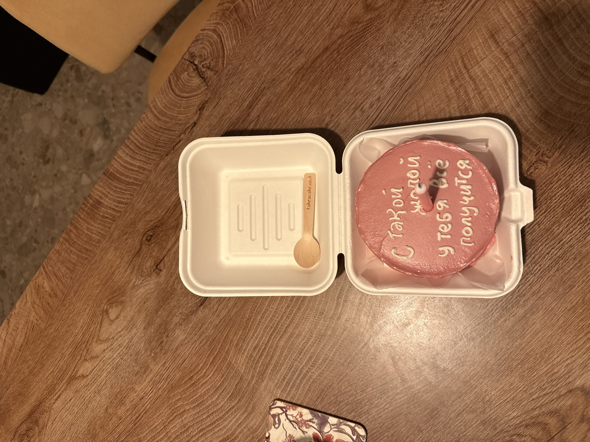 A small, round, pink cake is the central subject of the image, presented in an open white takeaway container on a wooden table. The cake is adorned with white icing text in Cyrillic script which reads, "С такой молодой у тебя всё получится," translating to "Being so young, everything will work out for you." A single, short white candle is inserted into the center of the cake.
Next to the cake, within the other half of the open container, rests a small wooden spoon. The spoon's handle is branded with "takeacake.co.il," a detail that ties the image to an Israeli context, specifically aligning with the provided Rishon LeZion location. The container itself appears to be a disposable, possibly eco-friendly, type.
The wooden table surface, showing distinct grain patterns, occupies most of the background and foreground. In the upper left, a blurry section of a light-colored, possibly beige or yellow, piece of furniture (likely a chair) is visible. A sliver of a patterned object, resembling a floral phone case or wallet, is partially visible in the lower foreground. The lighting is warm and ambient, suggesting an indoor setting, possibly during the evening. The scene evokes a personal, celebratory, or encouraging moment.