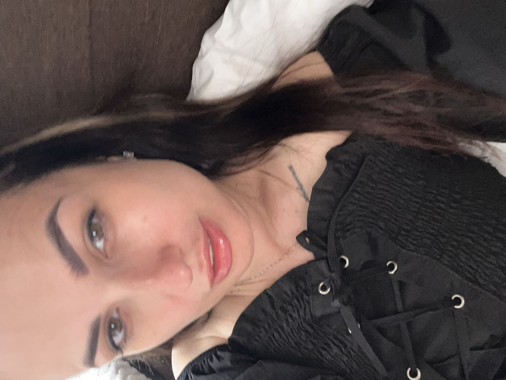 A woman is lying on a bed, facing the camera with her head slightly tilted. She has brown eyes, dark brown hair that falls over her shoulders, and is wearing a black, off-the-shoulder top with a lace-up front. The background is a blurred image of a headboard and a white pillow. The lighting suggests an indoor setting, possibly a bedroom. The woman's expression is neutral.