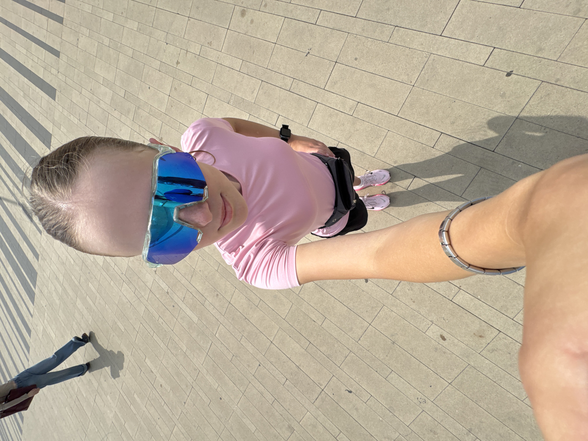 A young woman is taking a selfie outdoors on a paved surface. She is wearing a pink t-shirt, black shorts, pink sneakers, blue mirrored sunglasses, and a black fanny pack. A silver bracelet is visible on her left wrist. Her right arm is extended, holding the camera. In the background, another person wearing blue jeans and a dark maroon bag slung over their shoulder is walking away, casting a shadow. The ground is made of light-colored rectangular paving stones with darker striped shadows cast across them, suggesting an overhead structure or awning. The scene is illuminated by bright sunlight, creating distinct shadows. The setting appears to be an urban public space, possibly a walkway or plaza. Based on the user's request, this image could be located in Izhevsk, Russia.