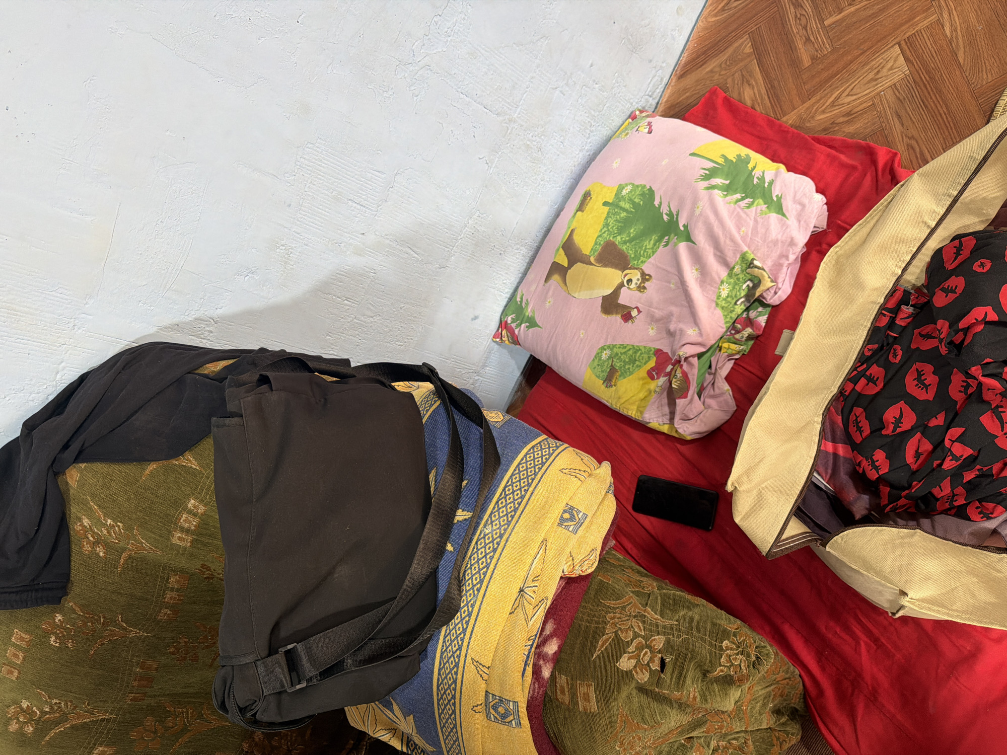 In Köhnə Quşçu, Azerbaijan, a flat lay image reveals an assortment of items arranged on a textured wall and wooden floor. A black messenger bag rests on top of a stacked pile of blankets. The top blanket is yellow with a blue patterned border, and beneath it is another blanket with a floral pattern in green and brown. A red blanket serves as a base for a pink pillow adorned with cartoon bears and green trees. A black smartphone lies on the red blanket, to the left of the pink pillow. A partially opened beige duffel bag sits on the red blanket, revealing black fabric with a red lip pattern inside. The scene is lit by overhead lighting, casting shadows on the textured wall and highlighting the details of the objects.