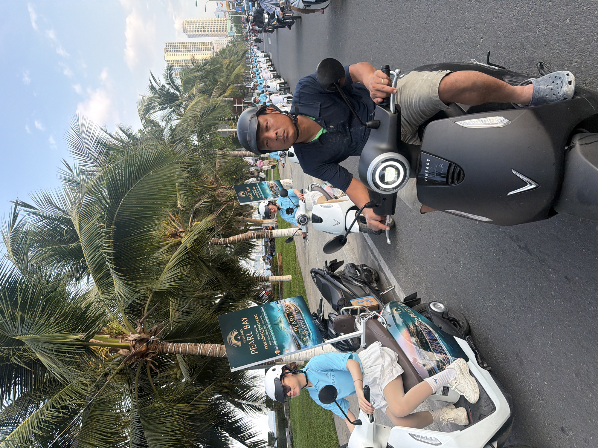 In Nha Trang Ward, Vietnam, a group of people ride electric scooters down a paved road lined with palm trees and tall buildings. The focus is on the lead scooter, ridden by a man wearing a black helmet and a dark blue shirt. Behind him, a woman in a light blue polo shirt and white skirt rides another scooter, displaying a "Pearl Bay" advertisement banner. Further down the road, a procession of similar scooters and riders stretches into the distance, suggesting a coordinated event or parade. The environment appears to be coastal, with a glimpse of the sea and sky visible behind the palm trees. The image captures a moment of organized movement within a bustling urban landscape.
