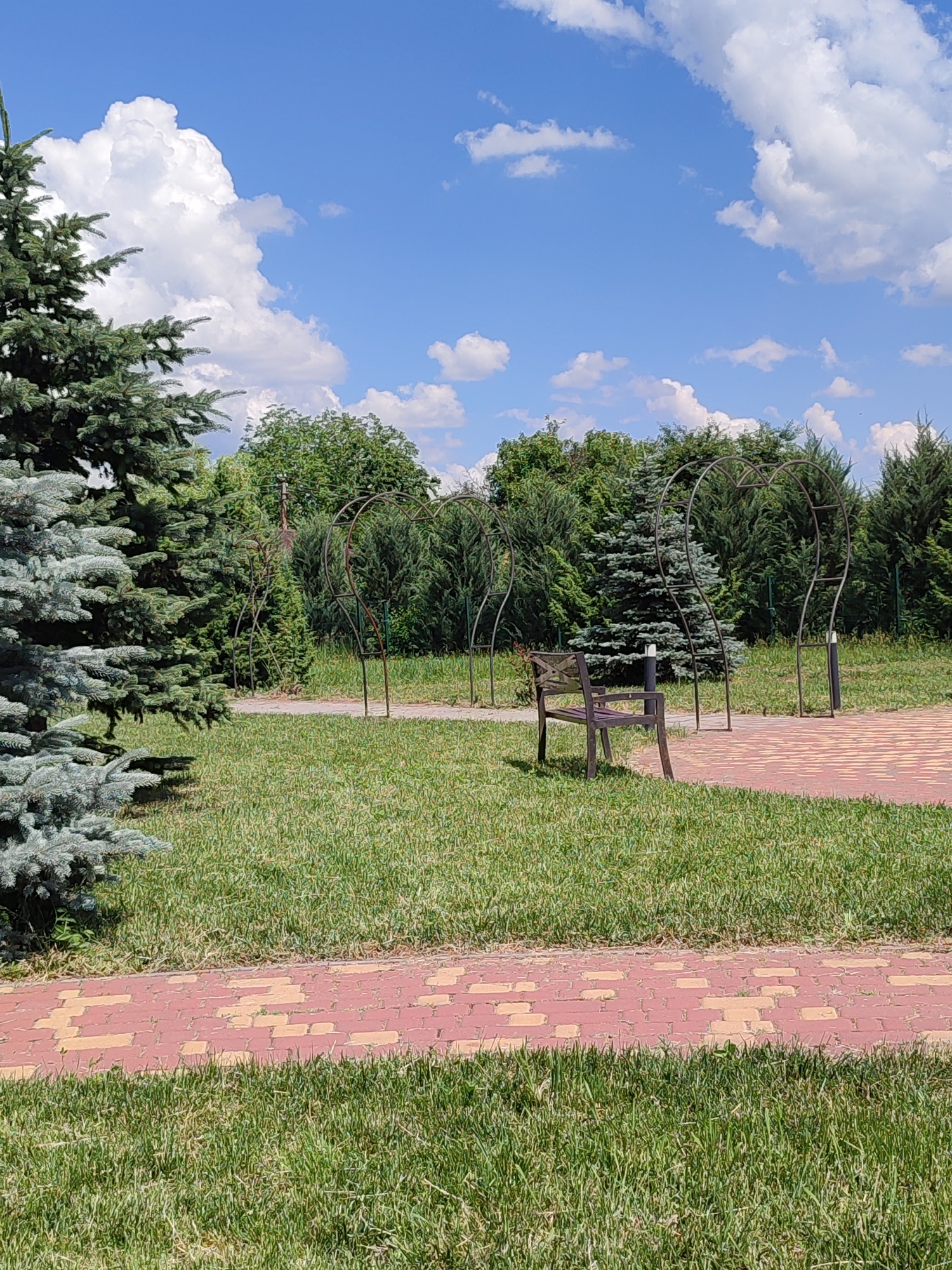 The image depicts a park scene on a clear, sunny day with a blue sky filled with scattered white clouds. In the foreground, a paved walkway made of red and yellow brick pavers runs horizontally across the frame, bordered by lush green grass. To the left, the lower branches of a large, blue-green spruce tree are visible.
Further into the park, the walkway continues, leading towards several heart-shaped metal trellises that appear to be decorative garden structures. A dark wooden bench sits on the grass to the right of the walkway, positioned in front of one of the trellises. Behind the trellises, a dense line of various green trees forms the background.
The overall environment suggests a well-maintained outdoor recreational area. The location is identified as the city of Voskobiinyky, Ukraine. There are no people or animals visible in the image.
