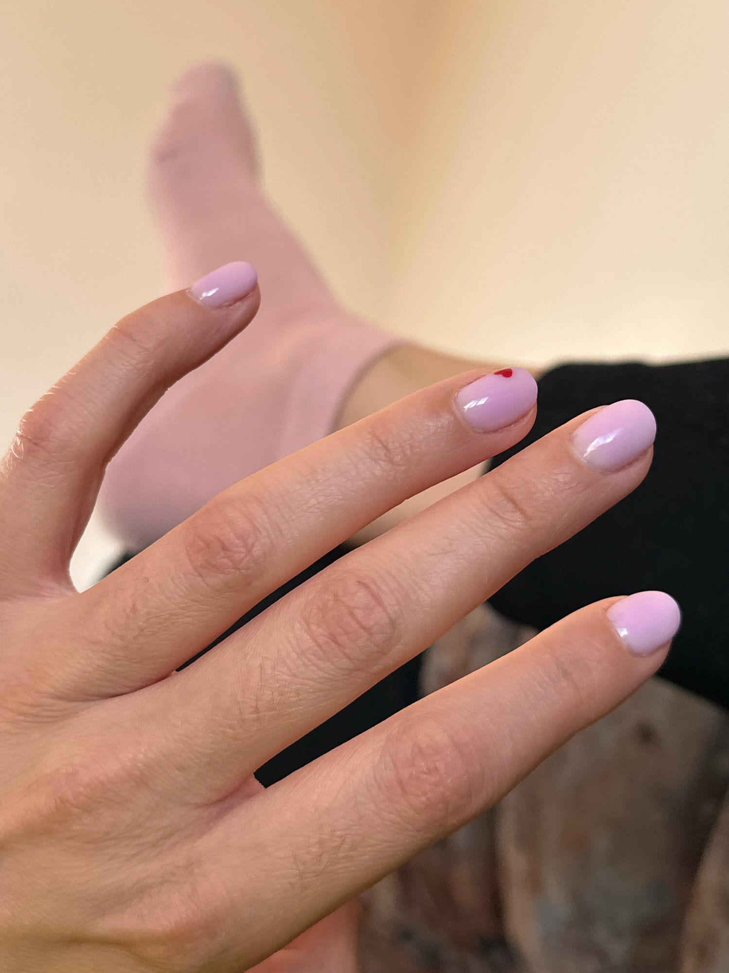 A close-up, high-angle shot focuses on a person's left hand and fingers, adorned with light purple nail polish. One fingernail, belonging to the index finger, features a small red heart design. In the background, a pale pink sock is visible, with the top portion of the leg beneath it. The environment appears to be indoors, with a neutral-colored background that transitions to darker tones, possibly indicating furniture or fabric. There is no discernible connection to the city of Gyumri, Armenia, in the provided image.
