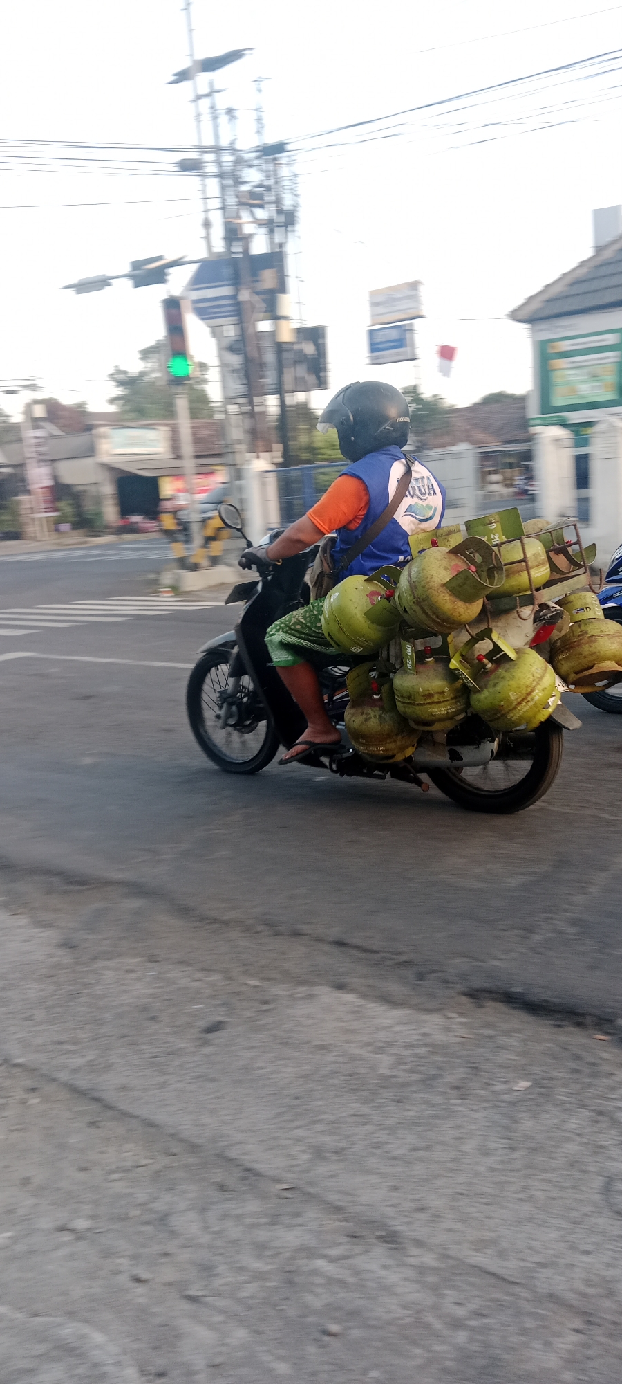 In Maguwoharjo, Indonesia, a person wearing a blue vest over an orange shirt and a black helmet rides a motorcycle laden with multiple green propane tanks. The motorcycle is traveling on a paved road, with a traffic light displaying a green signal in the background. Overhead wires and utility poles are visible, along with buildings and trees lining the street. The ground is asphalt, with some signs of wear and tear. The overall scene depicts a common mode of transportation and commerce in the area.