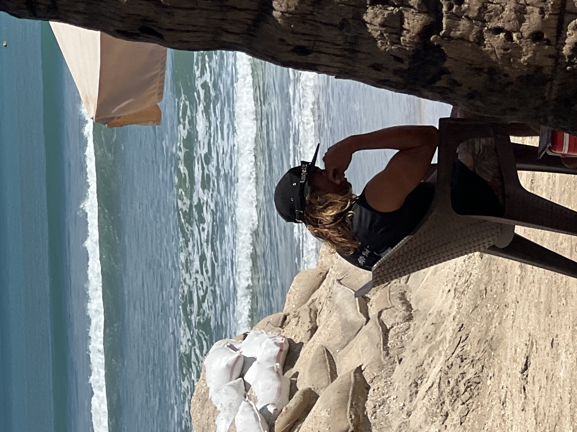 A person sits in a chair on a sandy beach, facing the ocean. The person is wearing a black t-shirt and a black baseball cap. They have long, light-colored hair and appear to be resting their chin on their hand. In the background, the ocean has moderate waves. To the left of the person, there is a pile of sandbags, some of which are white. To the right, a portion of a palm tree trunk is visible. A beige umbrella is partially visible behind the sandbags, suggesting a beachside setup. The overall scene depicts a relaxed atmosphere at the beach. This location is consistent with Kuta, Indonesia, a popular beach destination known for its surf and sandy shores.