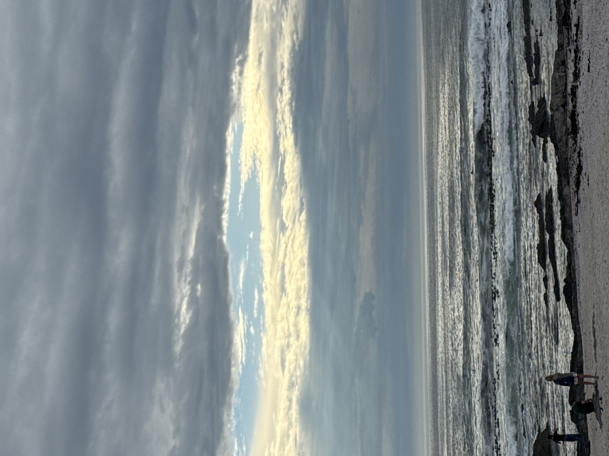 The image shows a seascape at Cape Town, South Africa. The sky is mostly cloudy with some patches of blue sky visible between the clouds. The ocean is slightly choppy, with waves breaking near the rocky shore. A few people are standing on the beach near the shoreline, possibly observing the ocean or interacting with each other. The beach appears sandy and slopes towards the water.