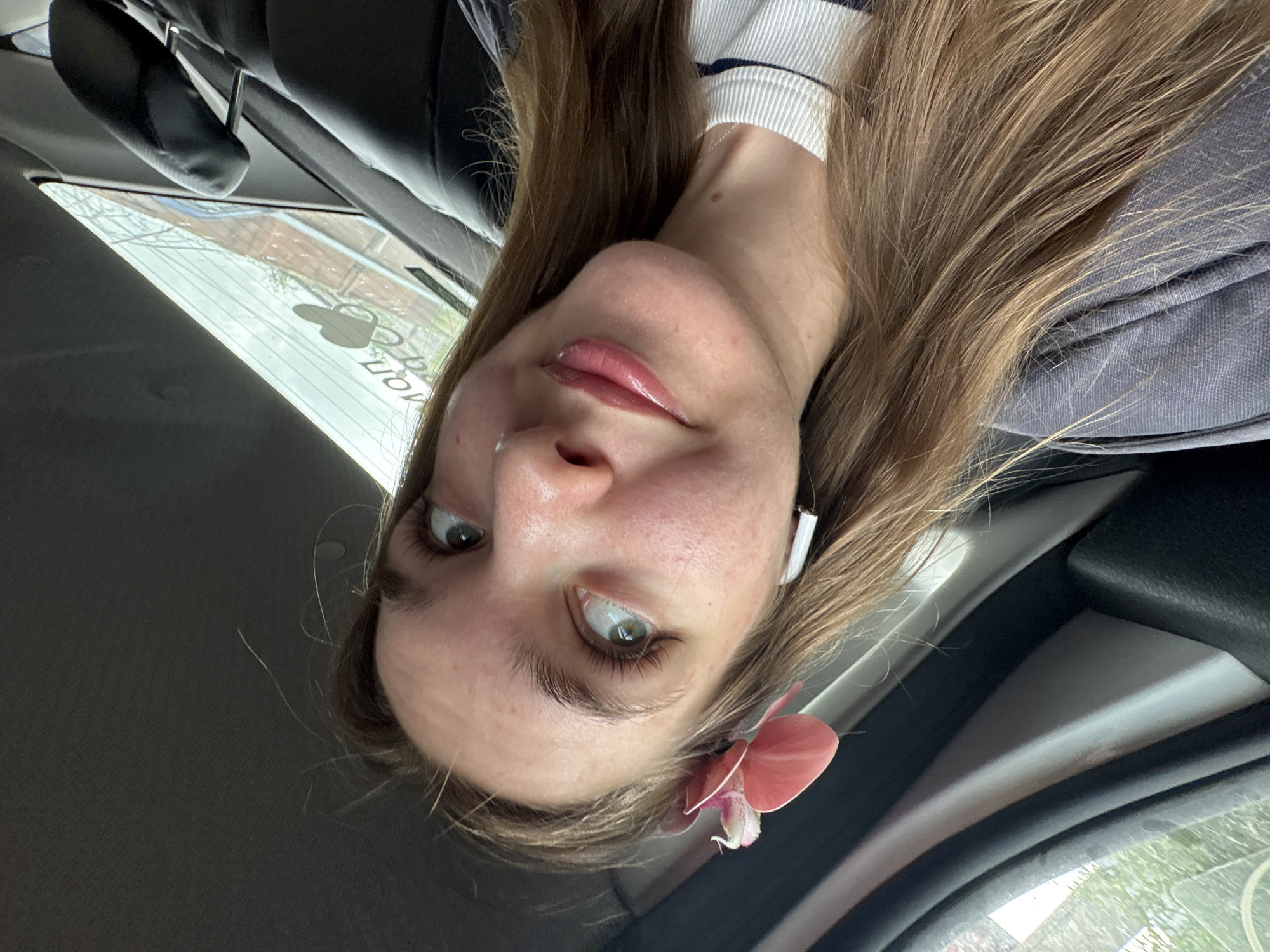 A young woman is depicted in a selfie taken from a low angle inside a vehicle. Her face is centrally positioned in the frame, with her head tilted slightly. She has fair skin, light brown hair, and light-colored eyes. She appears to be wearing a light-colored top with stripes and a grey jacket. There is a pink flower accessory attached to her hair near her ear. A white earbud is visible in her ear. The background shows a car interior, including the seats, windows, and other interior components. Through the car window, some outdoor scenery is visible, but it is out of focus and doesn't reveal clear details about Perm, Russia.