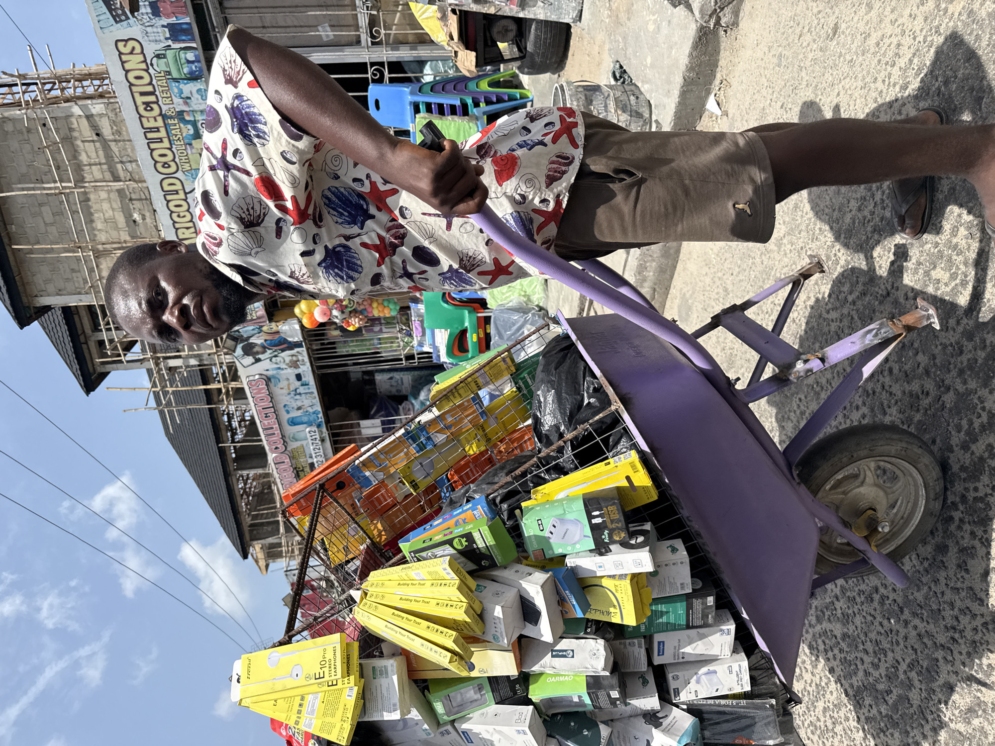 A man stands on a concrete sidewalk next to a purple wheelbarrow, its metal frame overfilled with packages of merchandise including earbuds and chargers, in Ado, Nigeria. The man wears a shirt patterned with red starfish and seashells, brown shorts, and sandals. Buildings and businesses are visible in the background, including one with the sign "RIGOLD COLLECTIONS WHOLESALE & RETAIL." The sky is blue and sunny.