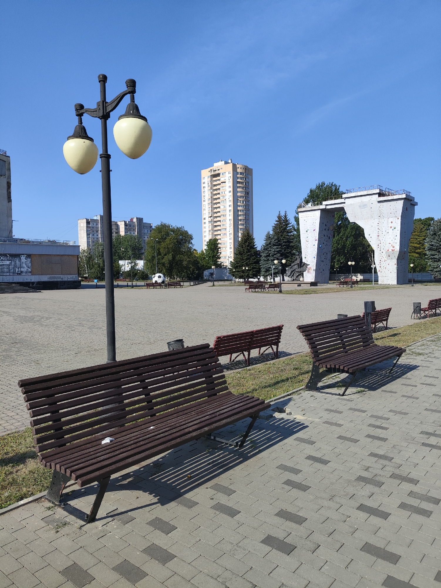 This image captures a sunny day in a public square in Kharkiv, Ukraine. In the foreground, two dark brown wooden benches with metal frames are placed on a paved area made of gray and dark gray rectangular tiles. The bench on the left is closer to the camera and casts long shadows. In the midground, a tall lamppost with two cream-colored, rounded lights stands prominently. Beyond the benches, several other benches and a trash can are visible. The square itself is mostly empty, with scattered green grass areas between the paved sections. In the background, a distinctive, large, white concrete structure resembling a climbing wall or an archway dominates the view, with climbing holds visible on its surface. To the left of this structure, a tall, slender, light-colored apartment building rises high into the clear blue sky. Other multi-story buildings and trees are scattered throughout the background, suggesting an urban environment. The overall scene conveys a sense of quietude and an outdoor recreational space.