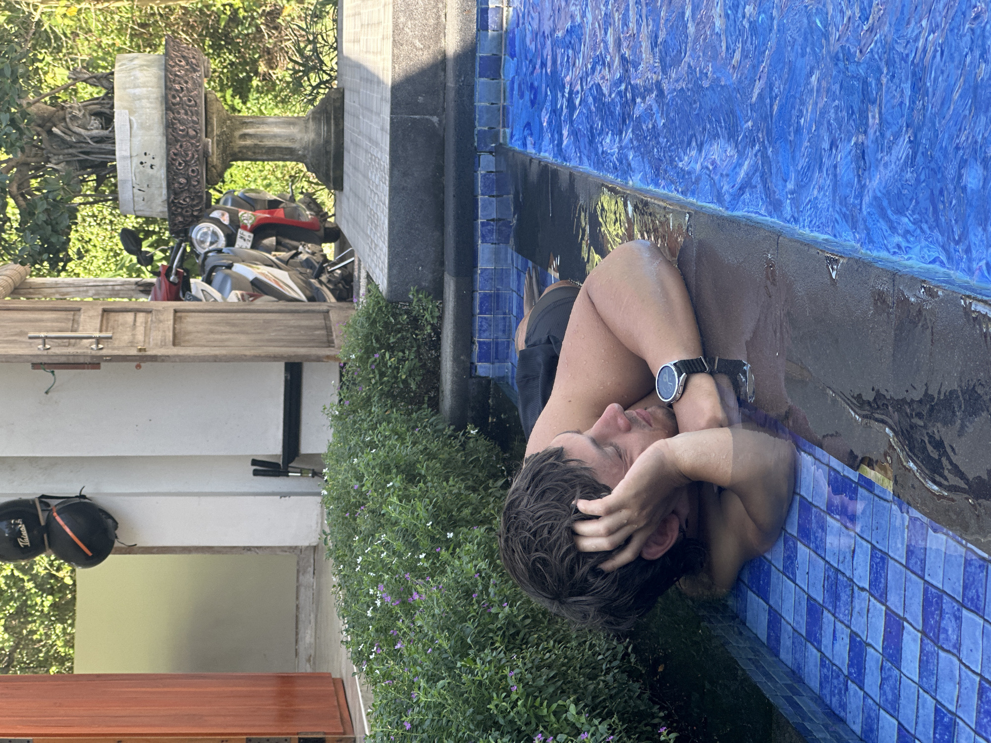 A man is in a swimming pool, with his head resting on the edge of the pool. He is wearing a wristwatch and appears to be relaxing. The pool is surrounded by greenery. In the background, there's a stone pillar or decorative structure, a building, and several motorbikes parked near a wooden gate or door. The location is likely in Nusa Penida, Indonesia. The pool has blue tiles, and the water is clear. The image is taken during daylight, with good lighting.