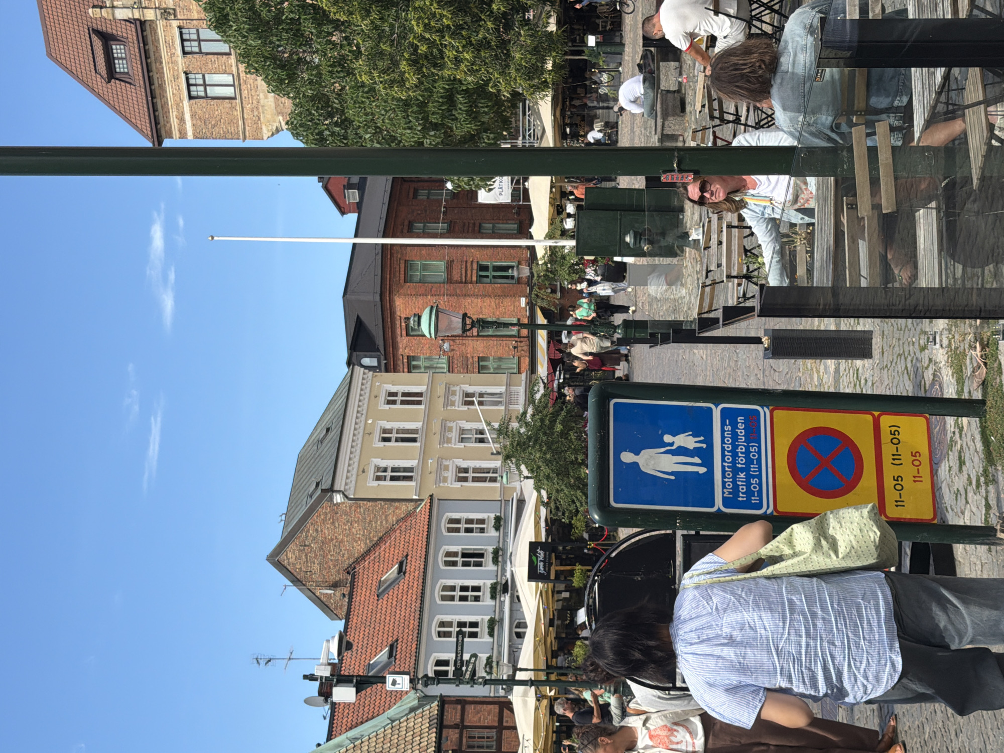 This is a medium-angle shot of a city square in Malmö, Sweden, likely Lilla Torg, on a bright, sunny day. The square is paved with cobblestones and features several outdoor seating areas for cafes and restaurants, indicated by tables and chairs. In the background, historic brick and stucco buildings with red-tiled roofs line the square. A tall, green lamppost stands prominently in the foreground, partially obscuring a view of the scene.
Several people are visible in the image. In the immediate foreground, a woman wearing a blue and white striped shirt and gray pants carries a light green tote bag over her left shoulder. She is facing away from the camera. To her right, a blue sign with white symbols indicates traffic restrictions. The top portion shows a pedestrian symbol, followed by text reading "Motorfordons-trafik förbjuden" (Motor vehicle traffic forbidden) and time restrictions "11-05 (11-05)". Below that, a circular red sign with a blue cross indicates no parking, with the same time restrictions "11-05 (11-05), 11-05".
Further back, several people are seated at outdoor tables. A woman with sunglasses and a light blue jacket is looking to her left. A person with long hair, wearing a denim jacket, is seated at another table. Other individuals are seen walking in the distance. A green awning with text in an unknown language is visible above a dining area. The sky is clear blue with a few wispy clouds.