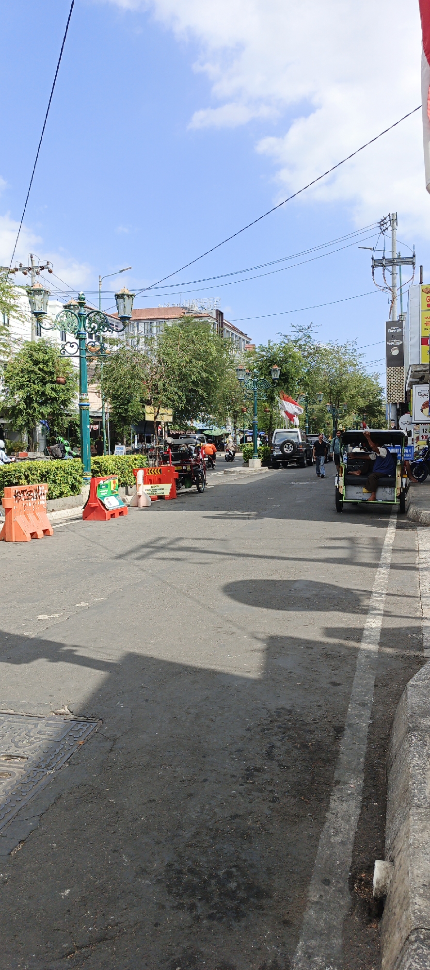 The image depicts a street scene in Yogyakarta, Indonesia, under a bright blue sky with scattered clouds. A paved road occupies the foreground and extends into the background, with a concrete curb and sidewalk visible on the right. Orange traffic barriers and some smaller white barriers are present on the left side of the road, with a green hedge behind them.
Several vehicles are on the road. In the immediate foreground, a shadow of a large object, possibly a tree or building, stretches across the asphalt. Further down the road, a three-wheeled vehicle, likely a bemo or becak motor, is stopped, with a passenger seated in the back. To the left of this vehicle, a red utility vehicle is parked. In the middle ground, a white SUV is parked. Several other motorcycles and a few people are visible in the distance, suggesting moderate activity.
The architecture in the background includes buildings with multiple stories, some with visible signage. Decorative street lamps, characteristic of older city streets, are present along the sidewalk. Lush green trees are interspersed with the buildings, providing shade and softening the urban landscape. An Indonesian flag is visible flying on a pole in the middle distance. The overall impression is of a busy urban street in a tropical climate, with a mix of traditional and modern elements.