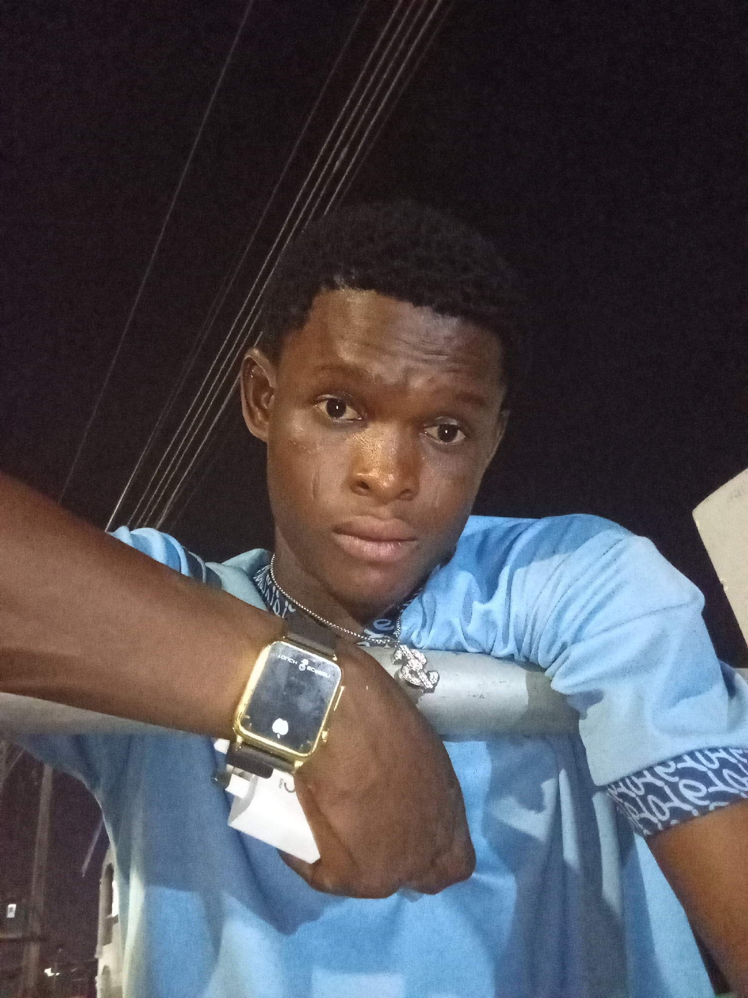 A close-up, eye-level portrait shows a young Black man leaning on a light-colored horizontal rail or pipe, looking directly at the camera. He is wearing a light blue short-sleeved shirt with patterned cuffs featuring a white design, and a silver-colored chain necklace with a dollar sign pendant. On his left wrist, he has a rectangular digital watch with a gold-colored frame and a black strap. A small white tag or paper is tucked under the watch strap. The watch face prominently displays "LORCH TECH" and a symbol resembling an apple logo.
The man has short, dark curly hair and dark eyes. His skin appears well-lit by an artificial light source, with highlights on his forehead, nose, and cheeks, suggesting a slight sheen, possibly from sweat. A faint mark or scar is visible on his left cheek (viewer's right). His expression is calm and somewhat serious.
The background is predominantly dark, indicating that the photo was taken at night. Thin, parallel white lines, likely power lines or cables, are visible in the upper and middle background. The setting is an urban outdoor environment, consistent with Ado, Nigeria.