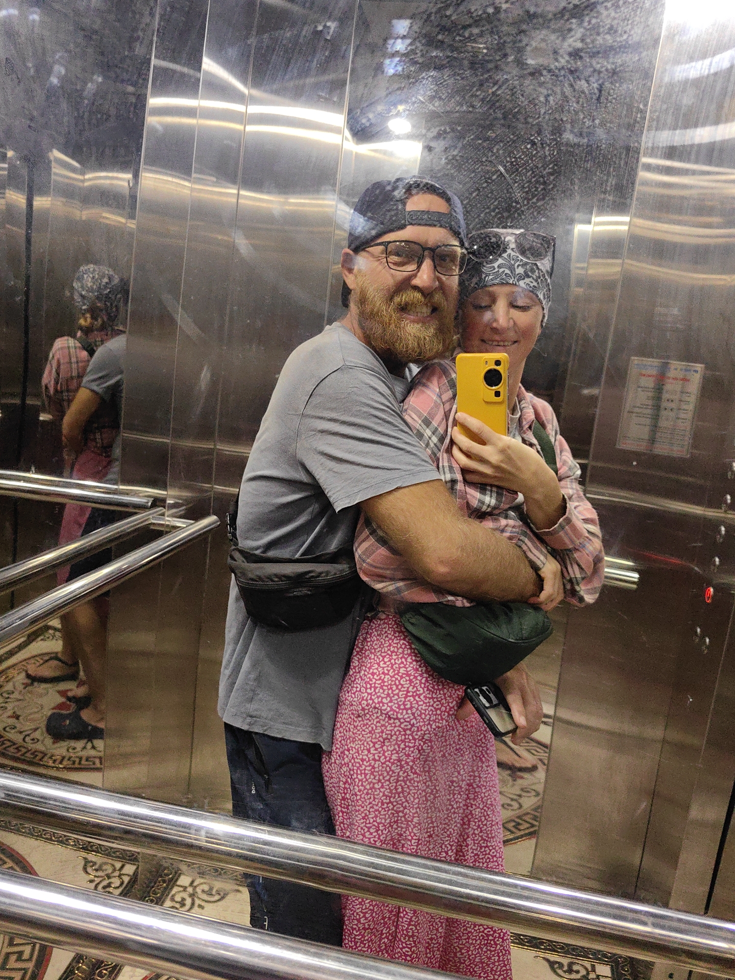 A couple is taking a selfie in an elevator, as indicated by the mirrored stainless steel walls and a chrome handrail visible in the foreground. The man is wearing a grey t-shirt, a baseball cap, and glasses, with a black fanny pack around his waist. He has a full beard. The woman is wearing a head covering, a plaid shirt, and a pink patterned skirt. She is holding a yellow smartphone. The floor of the elevator has an ornate patterned design. Information is not provided on any specific location in Bai Chay Ward, Vietnam.