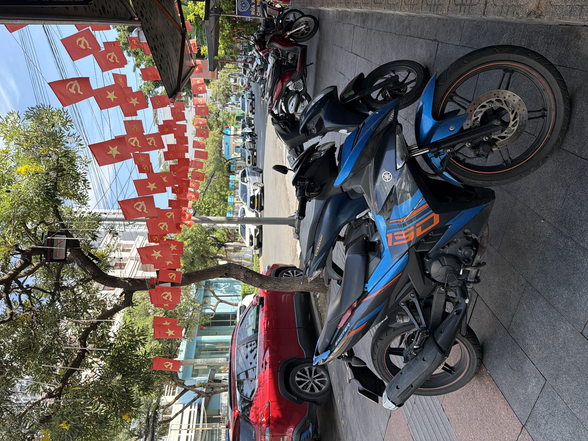The image depicts a street scene in Nha Trang, Vietnam, likely during a holiday or event. Red flags with yellow stars and hammers and sickles adorn overhead wires, stretching across the street. In the foreground, several parked motorcycles are visible, with a bright blue and black Yamaha sportbike being the most prominent. A red SUV is parked on the side of the road behind the motorcycles. The street is paved with gray and reddish-brown tiles, and a sidewalk runs along the right side of the image. Trees with green foliage and some yellow flowers are present, providing shade. Buildings line the background, and other vehicles, including cars and more motorcycles, are present on the street, suggesting a moderately busy urban environment. The sky is blue with some scattered clouds.