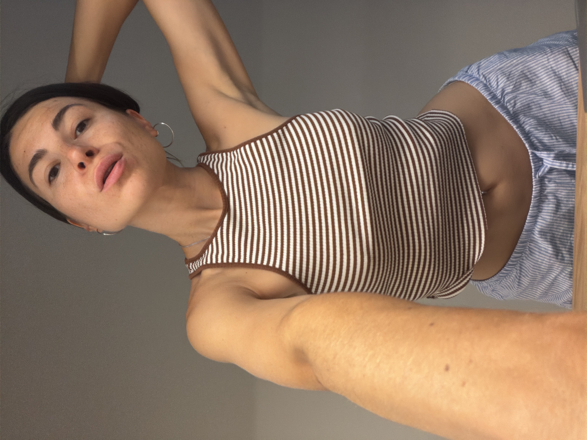 A woman with dark hair, wearing a brown and white striped top and blue and white striped shorts, is visible in a selfie-style image. She has light skin with freckles and is wearing hoop earrings. The background appears to be a plain gray wall. The location of the image within the city of Budva, Montenegro, is not discernible.