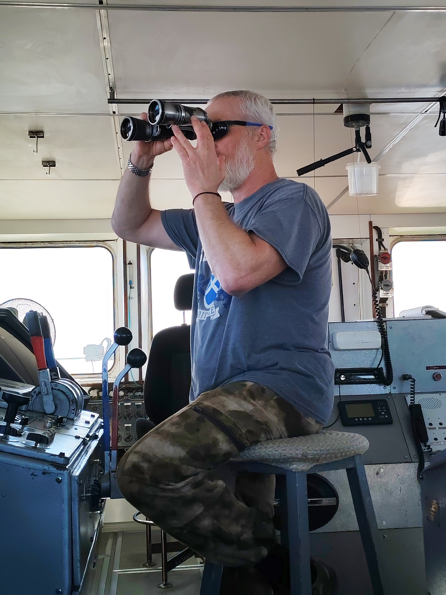 Inside a ship's bridge in Bol'shie Mkhi, Russia, a white male with gray hair and a gray beard is seated on a light-colored, round-padded stool. He is wearing dark sunglasses with blue temples, a dark blue short-sleeved t-shirt with a faint white graphic on the left chest, camouflage-patterned pants, a silver watch on his left wrist, and a black bracelet on his right wrist. He is holding a pair of black and silver binoculars to his eyes, looking forward and slightly upward.
The interior features light-colored walls and ceiling panels, with visible seams and overhead rails. Several metal hooks are suspended from the ceiling, and a white cylindrical device with a clear plastic bucket hangs from a rail on the upper right. Large, rectangular windows with bright white exteriors are visible on the left and right sides of the image.
To the man's left is a gray metal control console equipped with multiple levers, including prominent red and blue-handled ones, dials, and buttons. A circular metallic fan is partially visible behind this console. To his right, another gray metal panel incorporates various instruments, a small black screen labeled "VEGA," a black car-stereo-like unit, and a microphone on a coiled cable. Red and black control buttons are visible near the right window frame. A dark headrest of an additional seat is partially visible behind the man. The floor is light gray with visible seams.