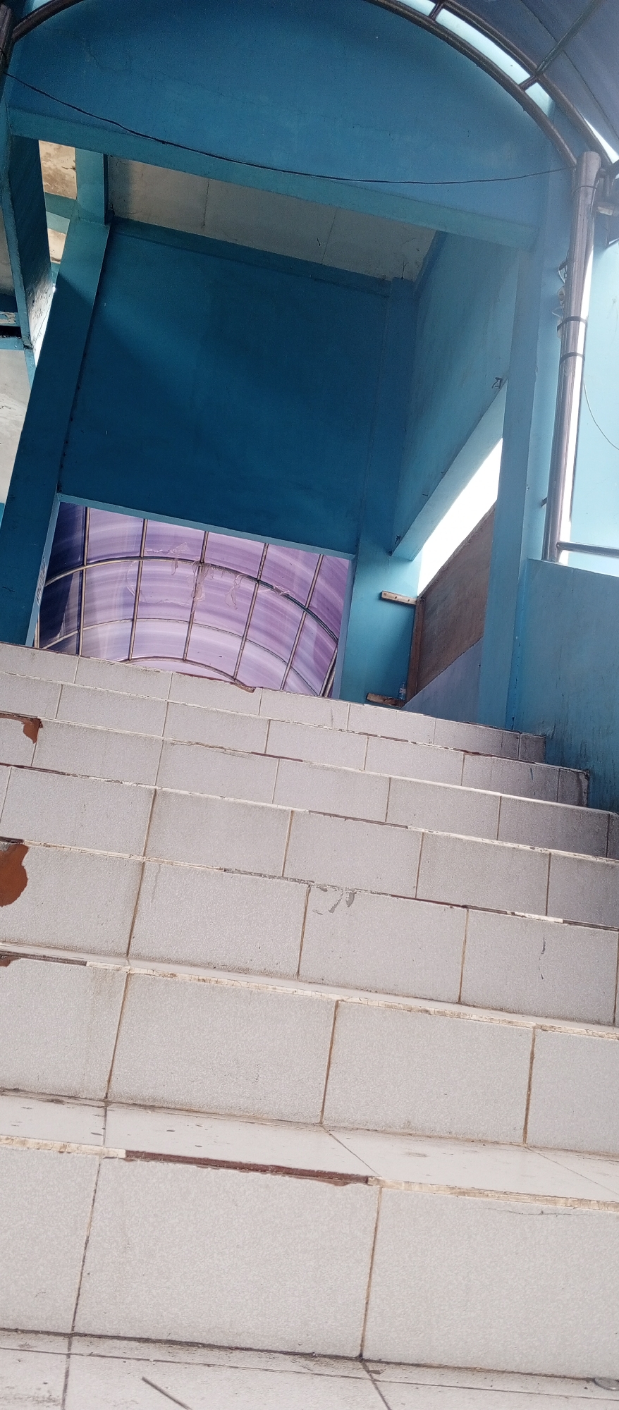 This is a ground-level, eye-level, vertical, medium shot of an outdoor staircase at the Plaosan Temple complex in Plaosan, Indonesia. The staircase is made of light-colored, square tiles and appears to be well-worn, with some chips and discoloration on the edges. The stairs lead upwards towards a bright, open space illuminated by sunlight. The walls of the staircase structure are painted a vibrant blue. Above the staircase, a translucent purple roof with a grid pattern is visible, supported by a curved metal frame. The overall scene suggests a functional outdoor space within a historical or cultural site, characterized by contrasting colors and textures.
