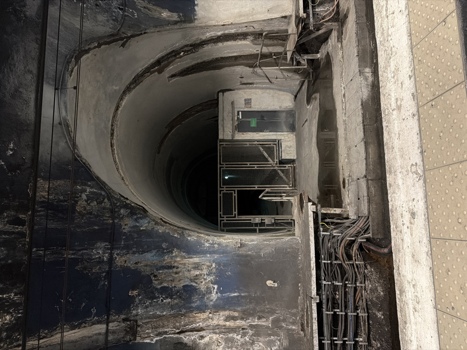 The image displays a dimly lit, static scene within an underground tunnel section, identified as likely part of the Paris Métro system. In the foreground, the edge of a light-colored platform featuring tactile paving is visible. Immediately beyond this, a concrete ledge runs parallel to the platform, beneath which numerous bundled electrical cables and conduits are exposed along the wall. The overhead tunnel arch exhibits significant water damage, characterized by large patches of dark blue, black, white, and brown discoloration, consistent with efflorescence and grime.
In the midground, a metal-grated entrance, partially ajar, leads into a darker, deeper section of the tunnel, with a faint light source visible within. The uneven concrete floor directly in front of this gated entrance features a substantial puddle of water. Adjacent to this, to the right, a dark grey door with a green sign is recessed into a concrete wall. Further to the right, a raised concrete platform with visible wooden supports and exposed cables underneath includes a metal handrail ascending from the right side. Another, smaller puddle of water is present on the concrete floor in this area. No individuals or active subjects are present within the scene.